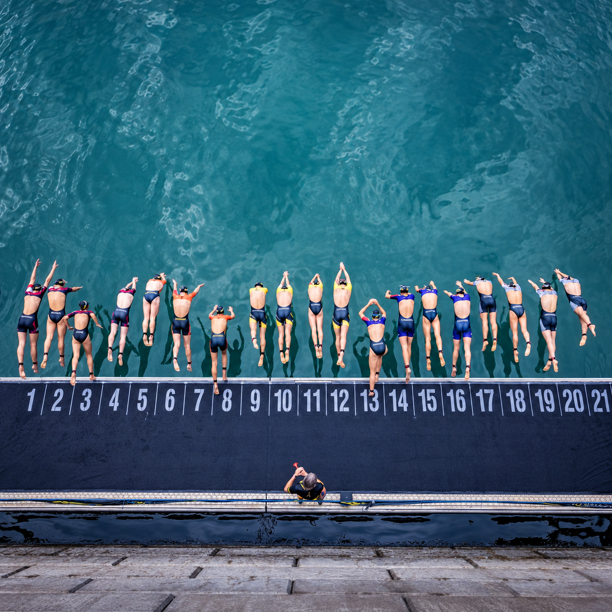 A group of synchronized swimmers in swimsuits and swim caps, lined up on a diving platform before a race, with their shadows cast on the water below.