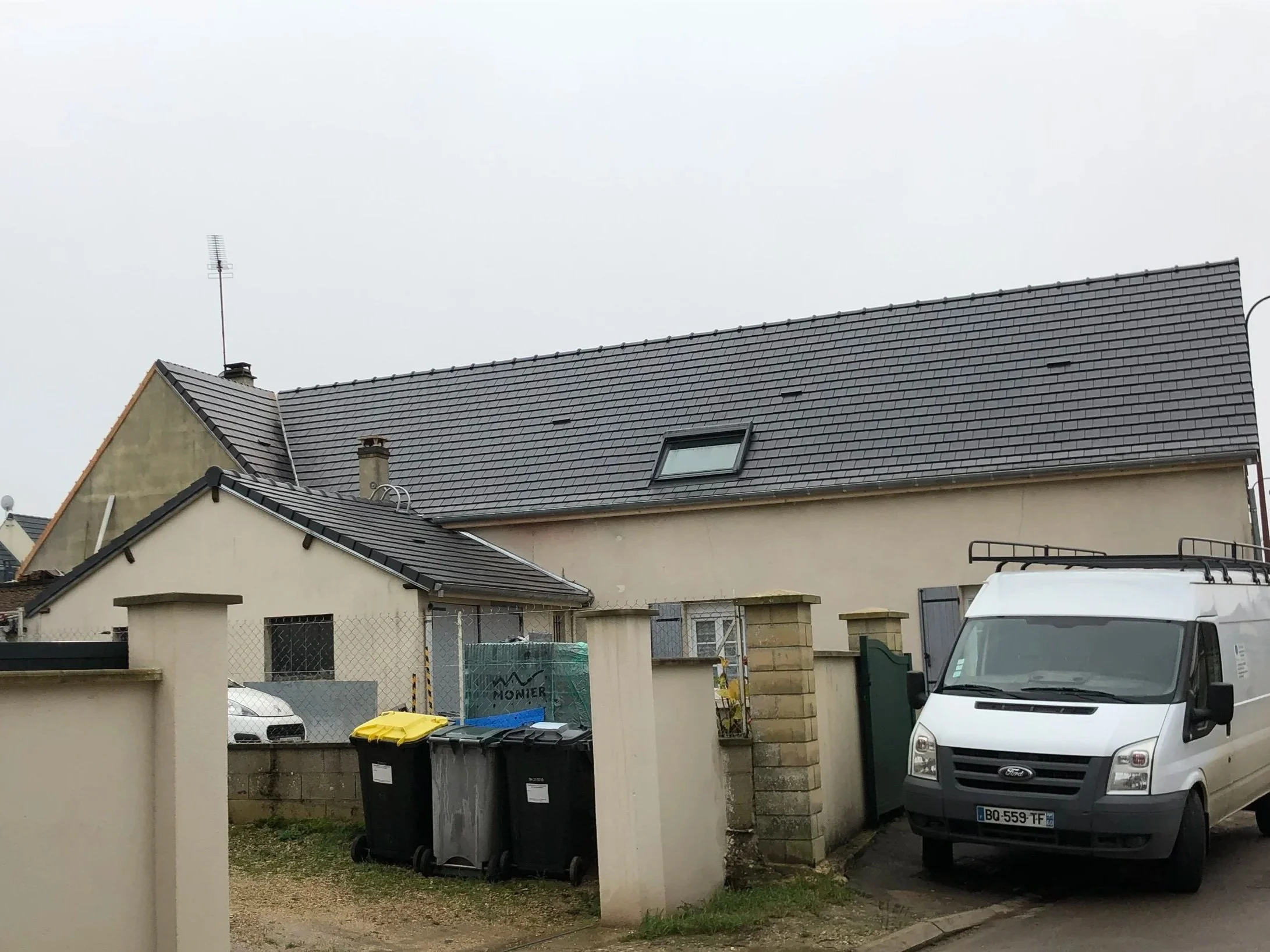 Maisons avec toits en tuiles grises, voiture blanche, poubelles noires et jaunes, clôture en métal, ciel nuageux.