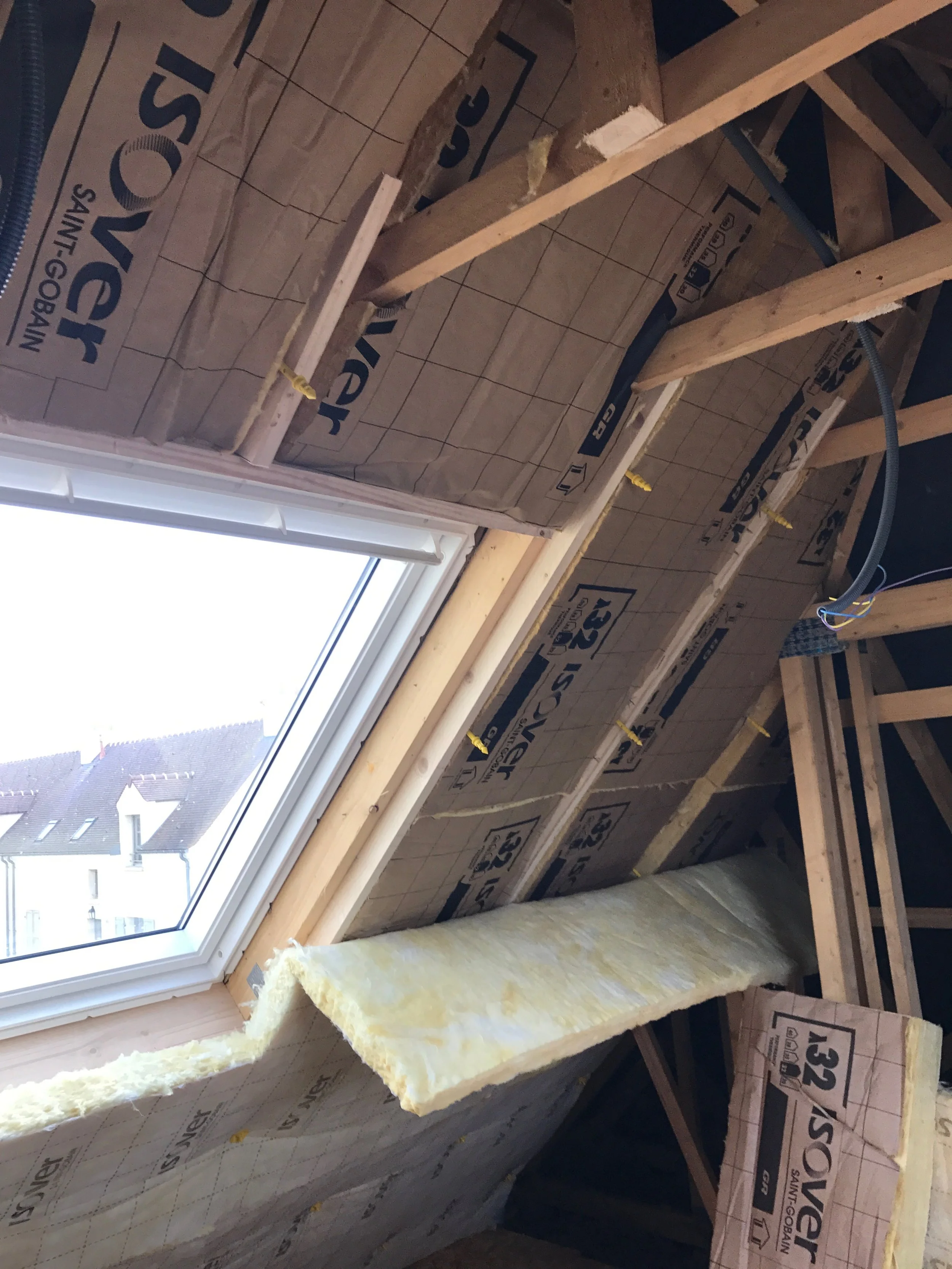 Travaux d'isolation dans une toiture avec un velux, des murs en construction et des câbles électriques visibles.
