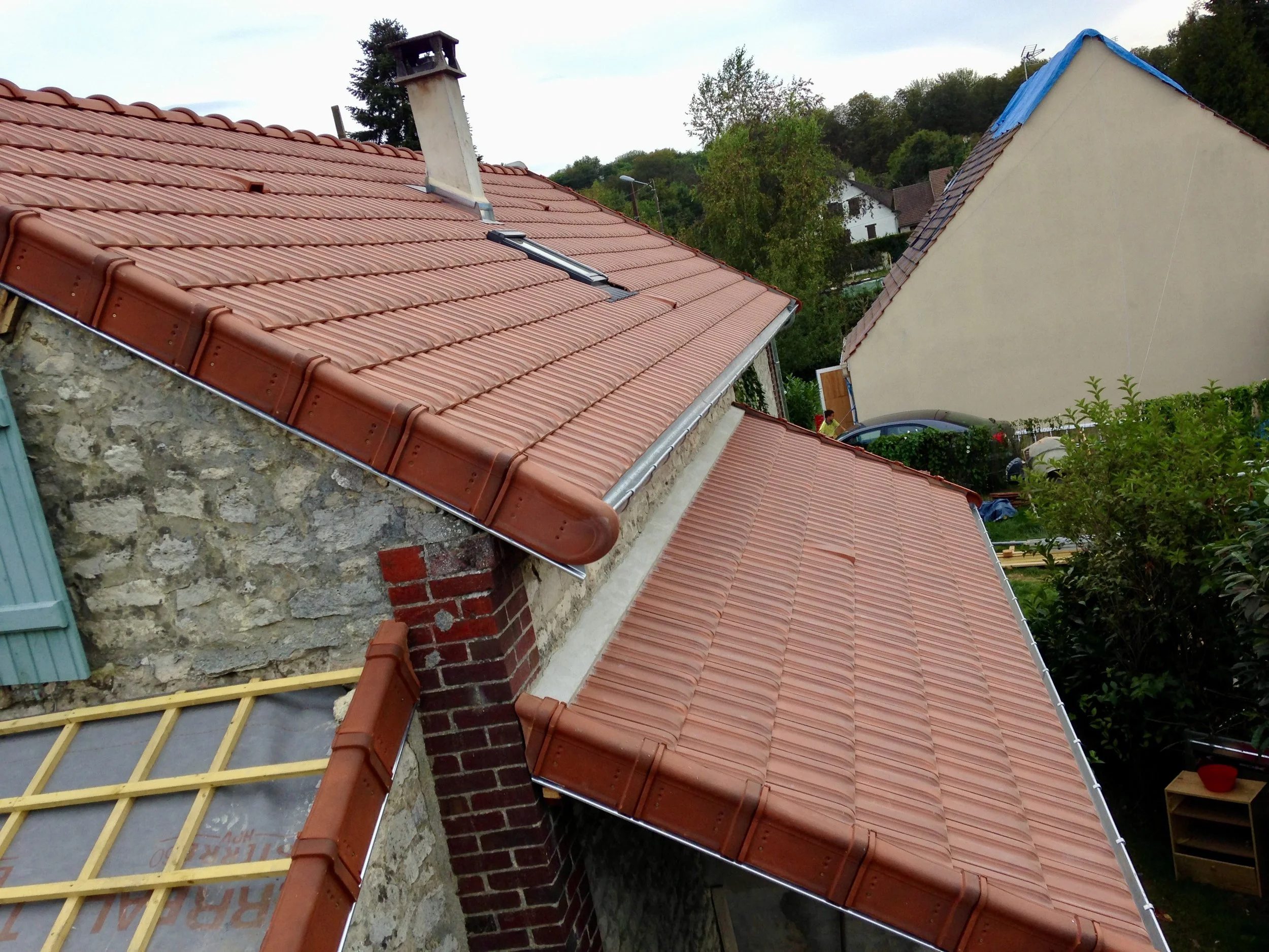 Transformation de charpente d'une maison en pierre avec fenêtres et toit en pente, arbres et autres maisons en arrière-plan.
