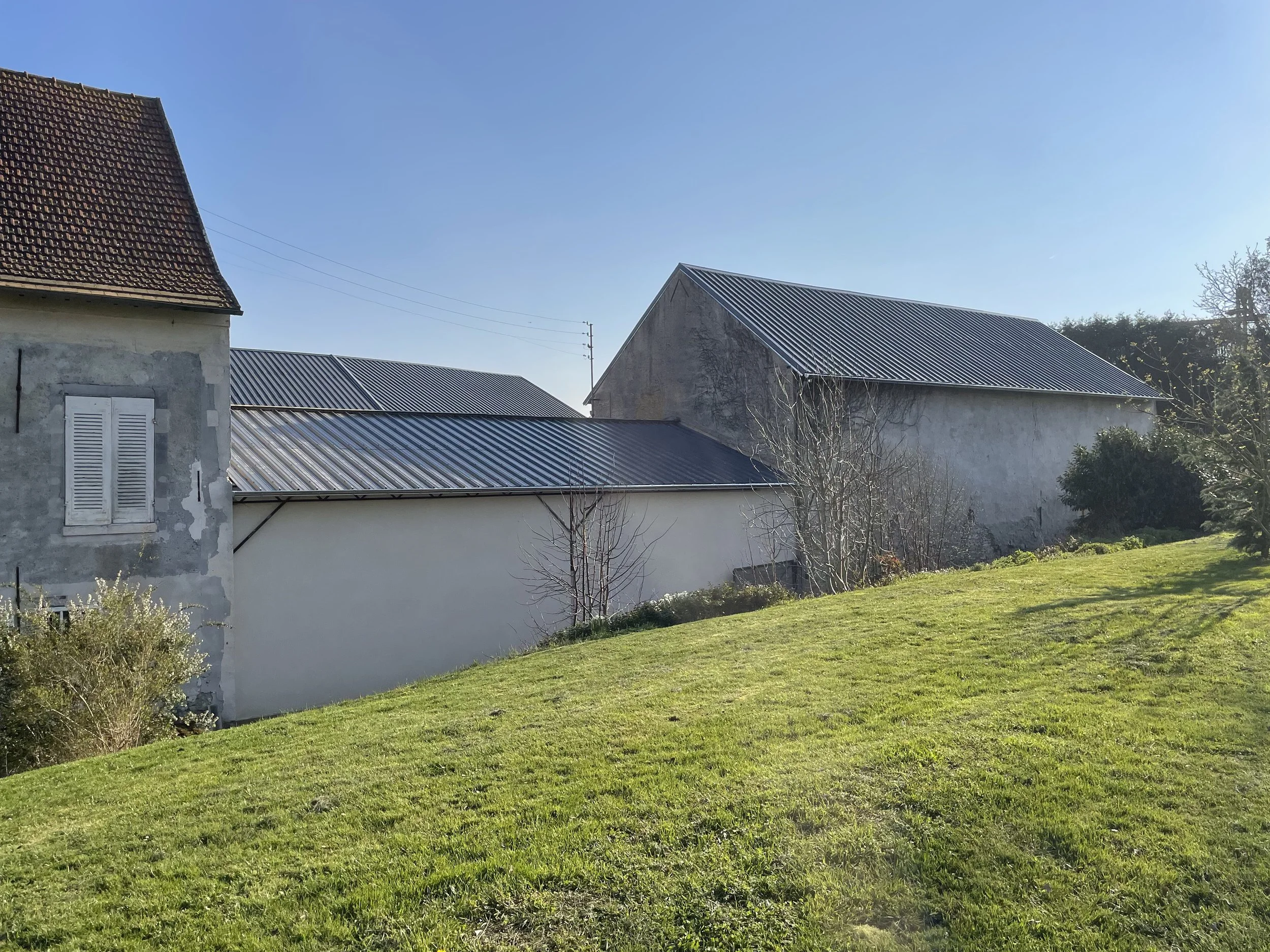 Maisons anciennes en pierre avec toitures en tuiles et en métal dans un paysage ensoleillé avec un grand espace vert et des arbres, ciel clair sans nuages.