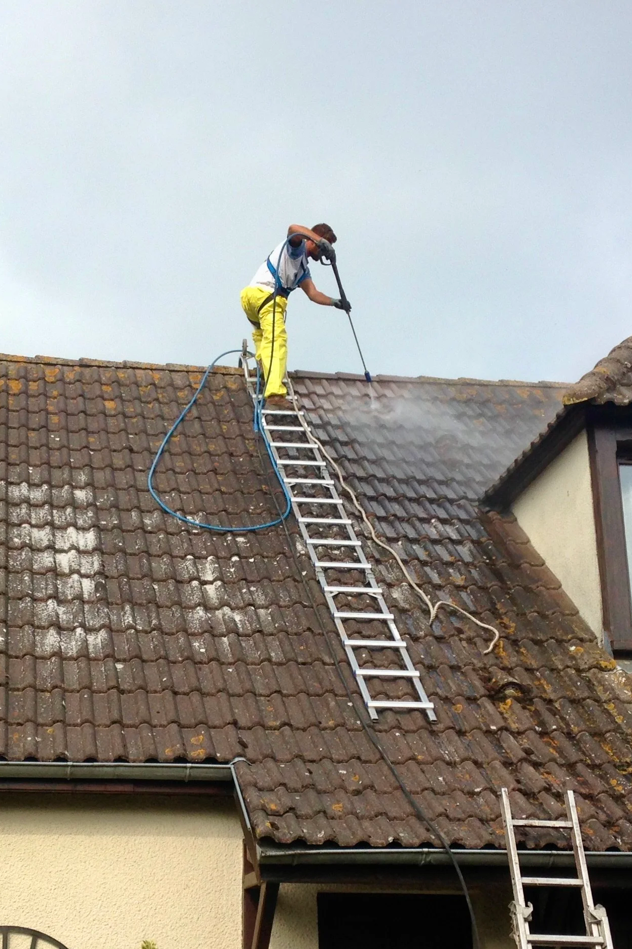 Un homme en vêtements de travail jaune entretien la toiture d'une maison avec un hydrofuge coloré en utilisant une échelle pour accéder au toit.