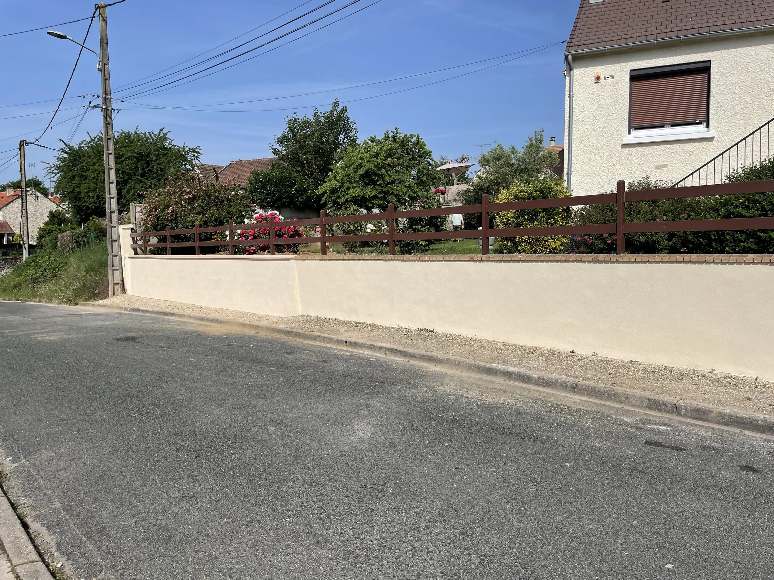 Vue d'une rue résidentielle avec une clôture en bois, des arbres en fleurs, une maison avec fenêtre à volet roulant fermé, des câbles électriques suspendus, sous un ciel bleu clair.
