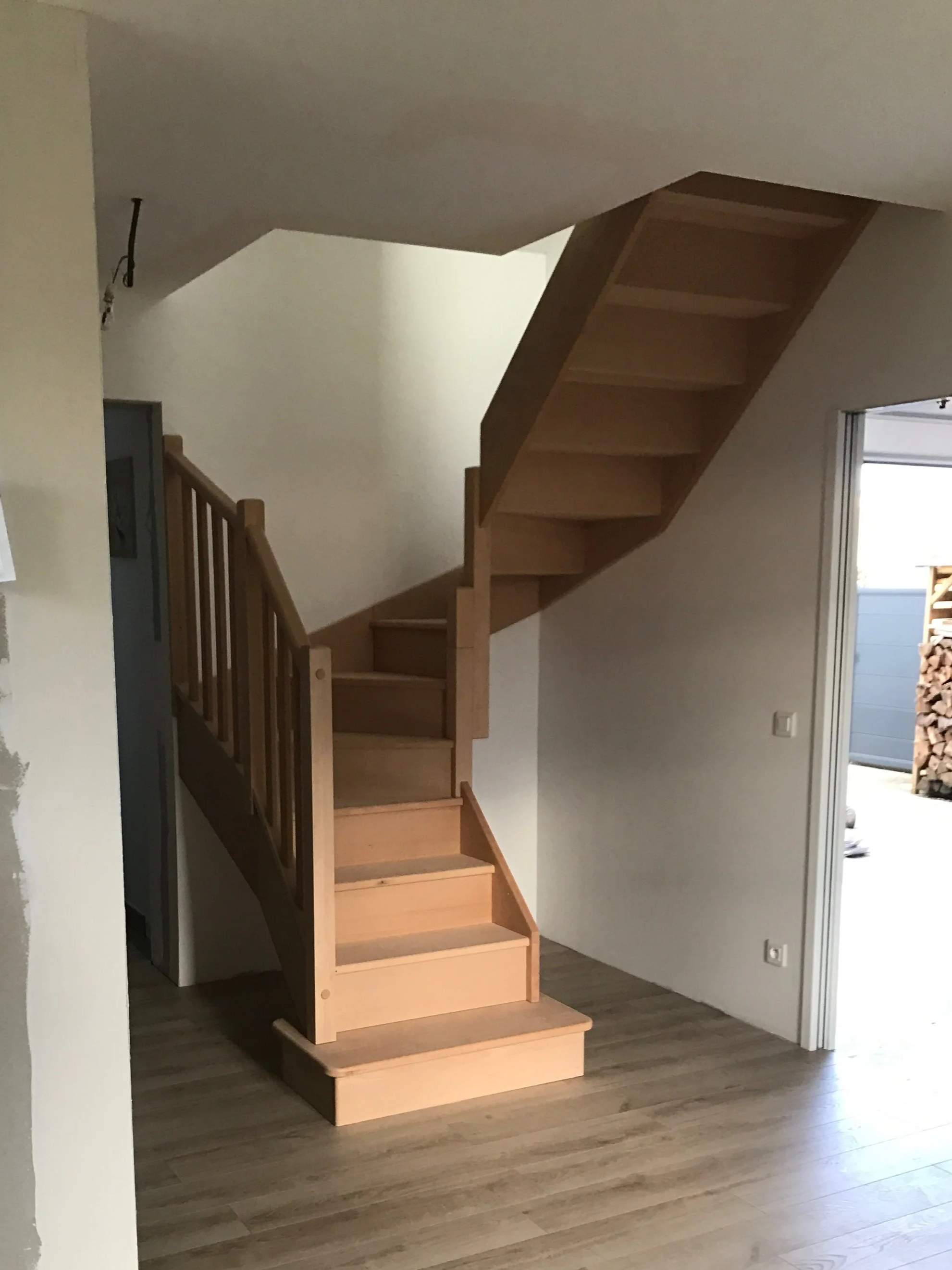 Escalier en bois intérieur avec marches en bois clair et rampe en bois, dans une pièce avec sol en bois et mur blanc.