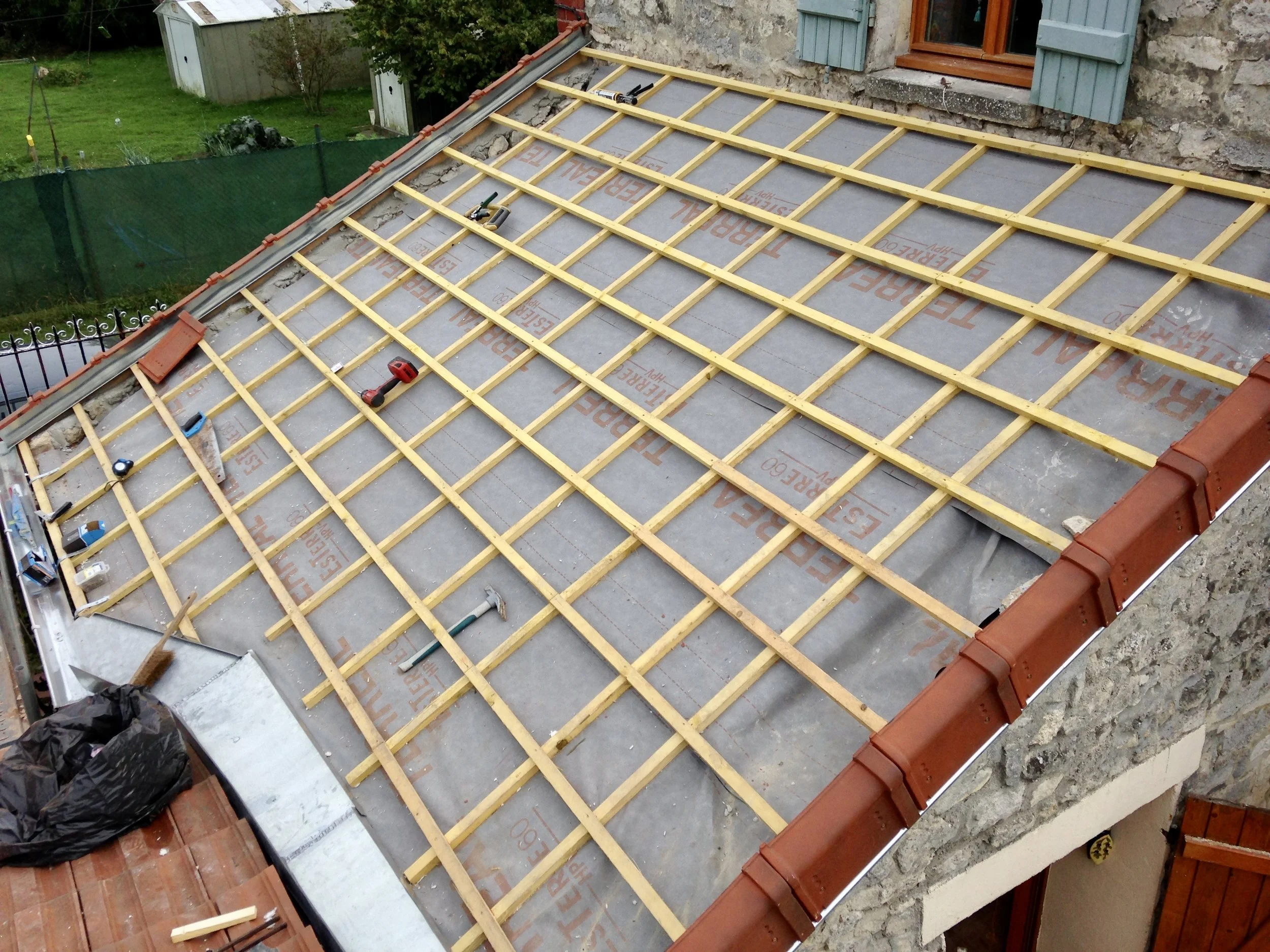 Transformation de charpente d'une maison, avec des outils posés dessus, entourée d'une maison en pierre et d'un jardin vert avec un abri et des arbres.