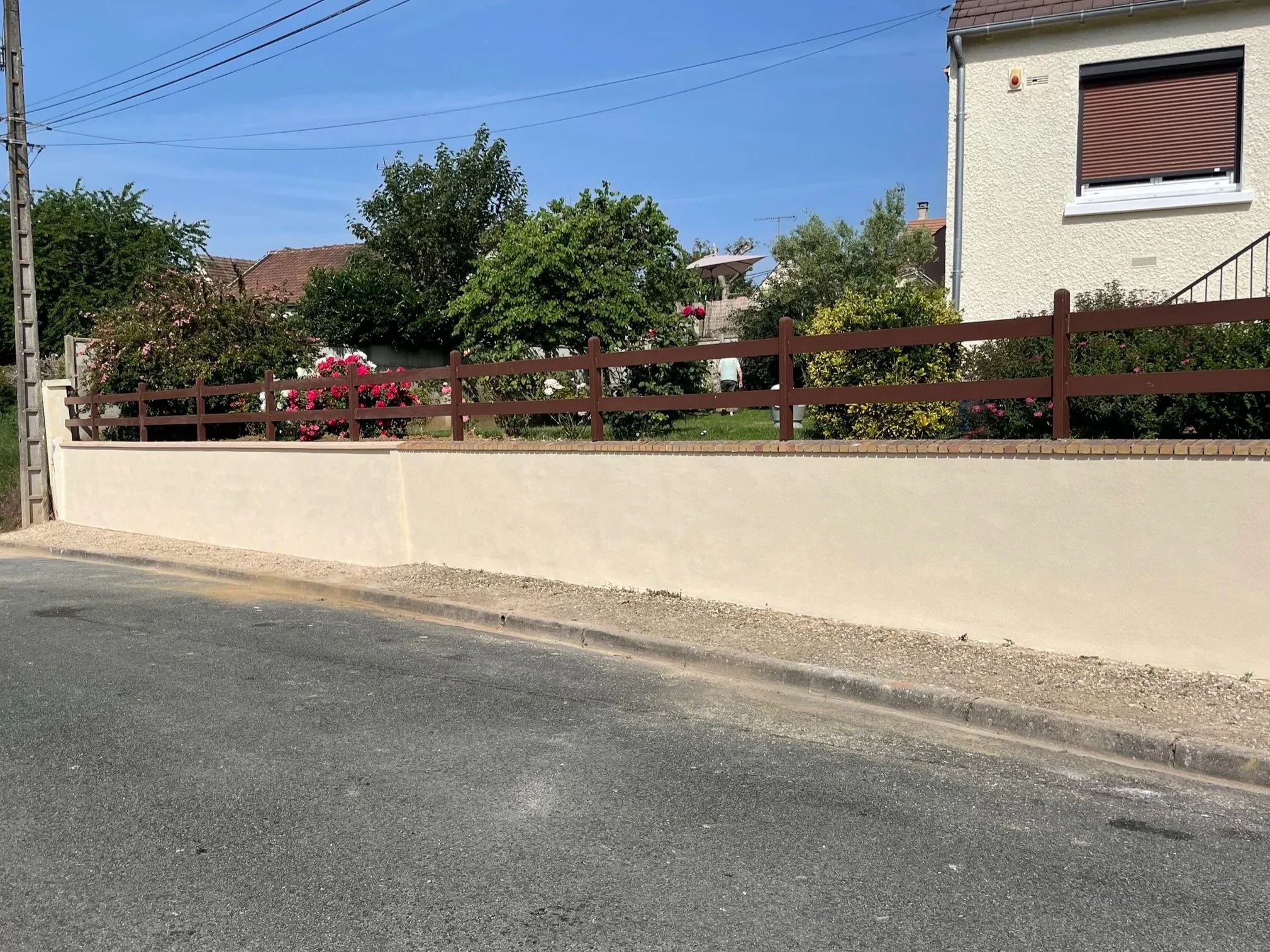 Une vue d'un mur maçonné fini, une clôture en bois, des arbres et une maison en arrière-plan sous un ciel bleu.