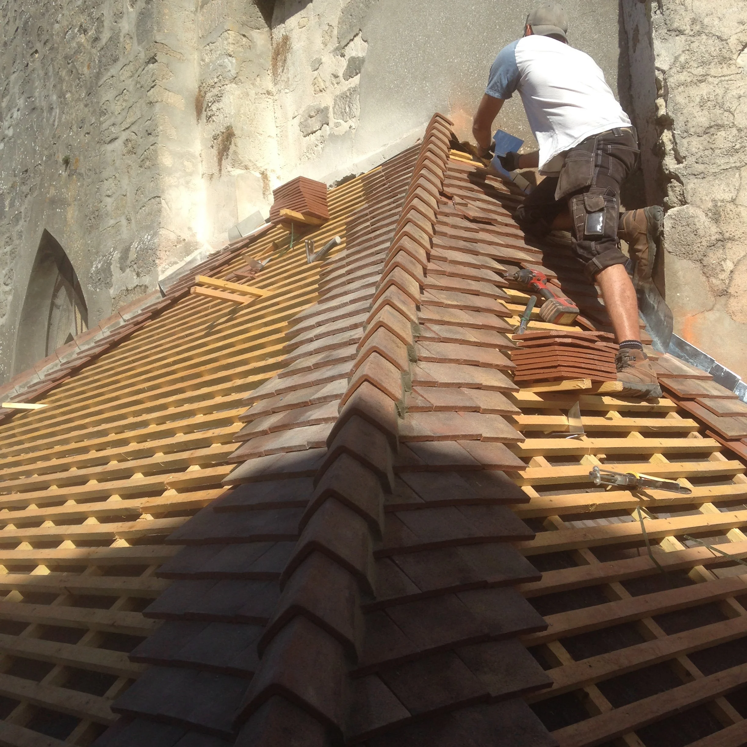 Un homme construit ou répare une couverture en tuiles en terre cuite sur un toit en utilisant des outils, avec un mur en pierre en arrière-plan.