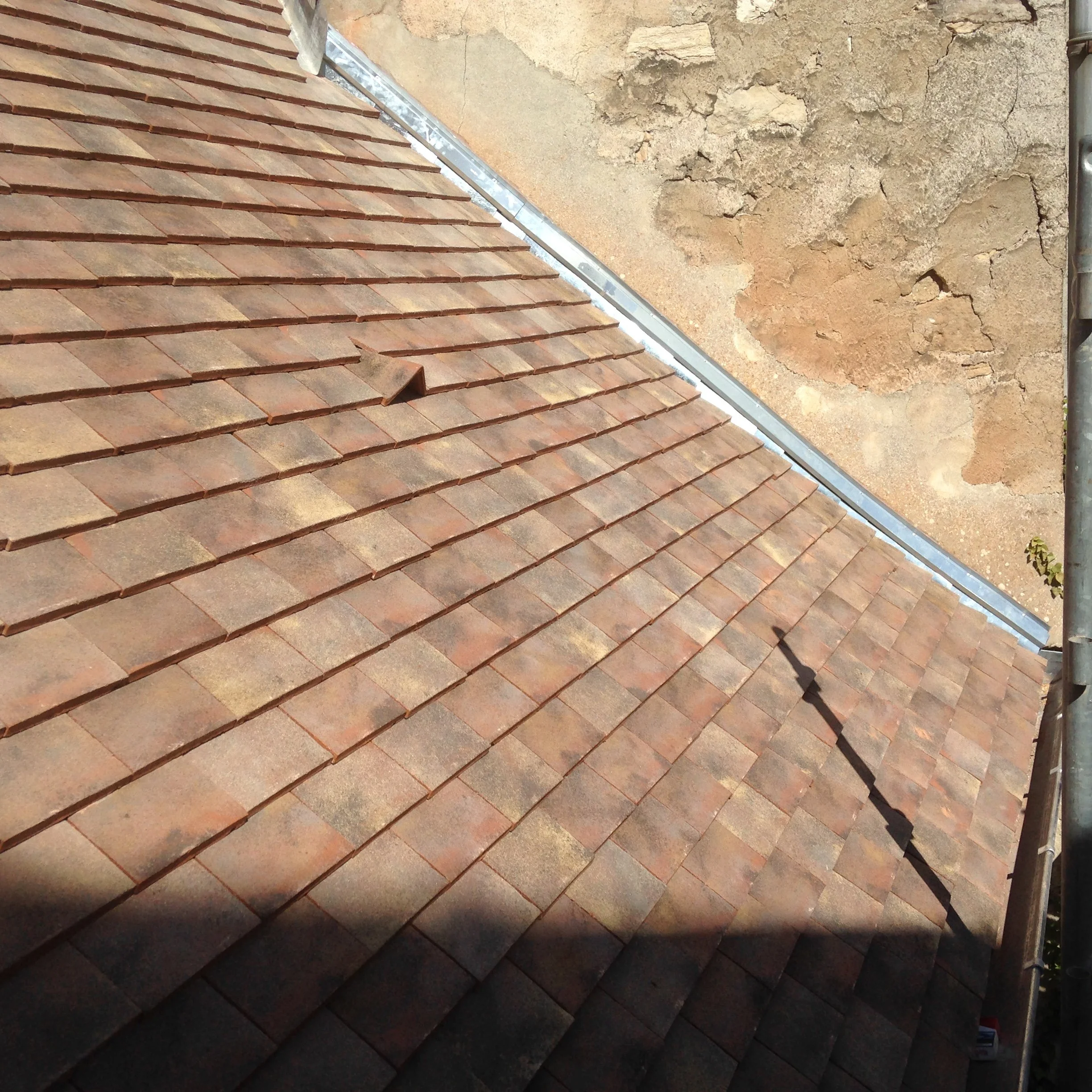 Vue partielle d'une toiture en tuiles rouges avec une gouttière en métal, et un mur en pierre avec des fissures.