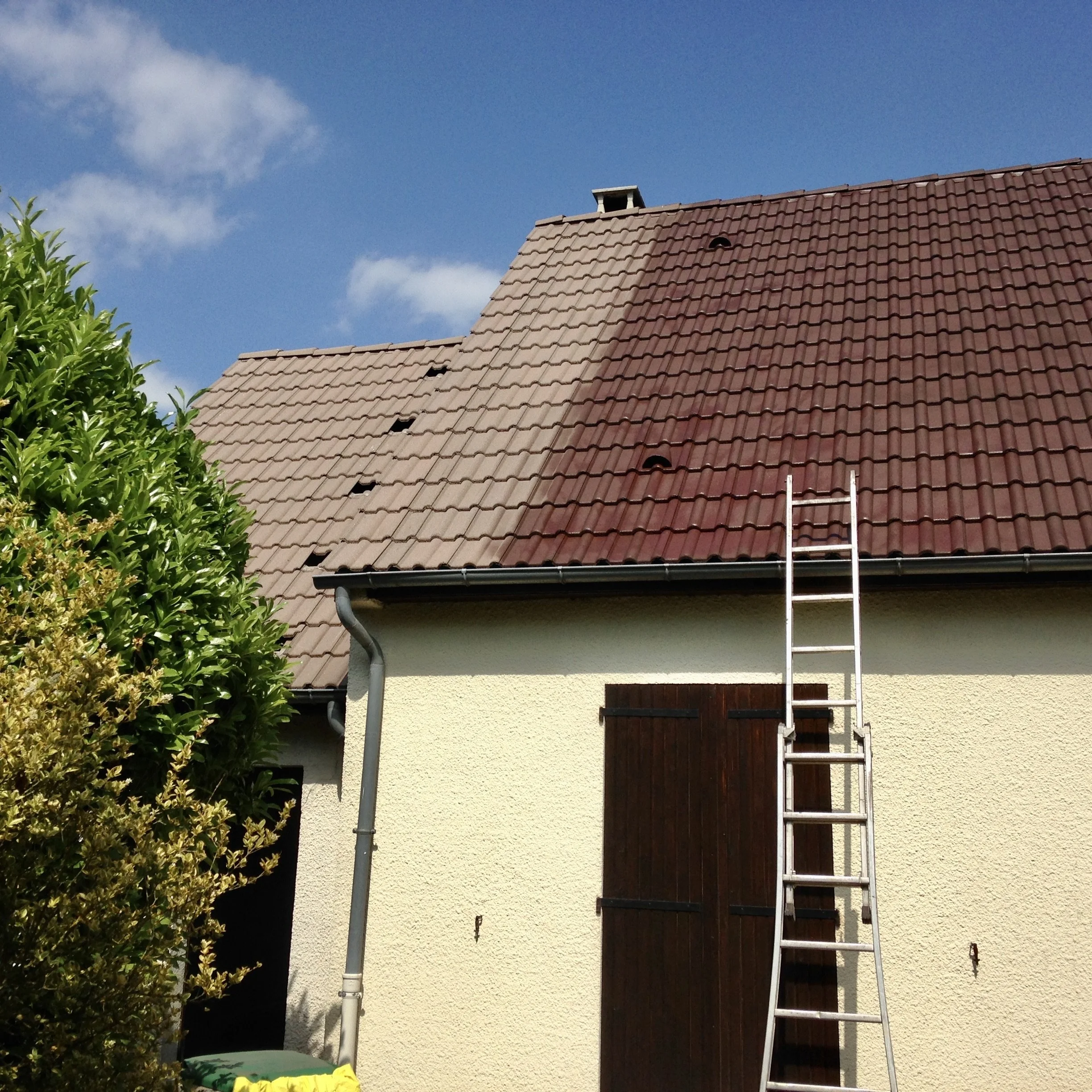 Toit de maison en tuiles rouges, cheminée, fenêtre de toit, escabeau en aluminium, gouttière, mur beige, végétation verte de gauche, ciel bleu avec quelques nuages.