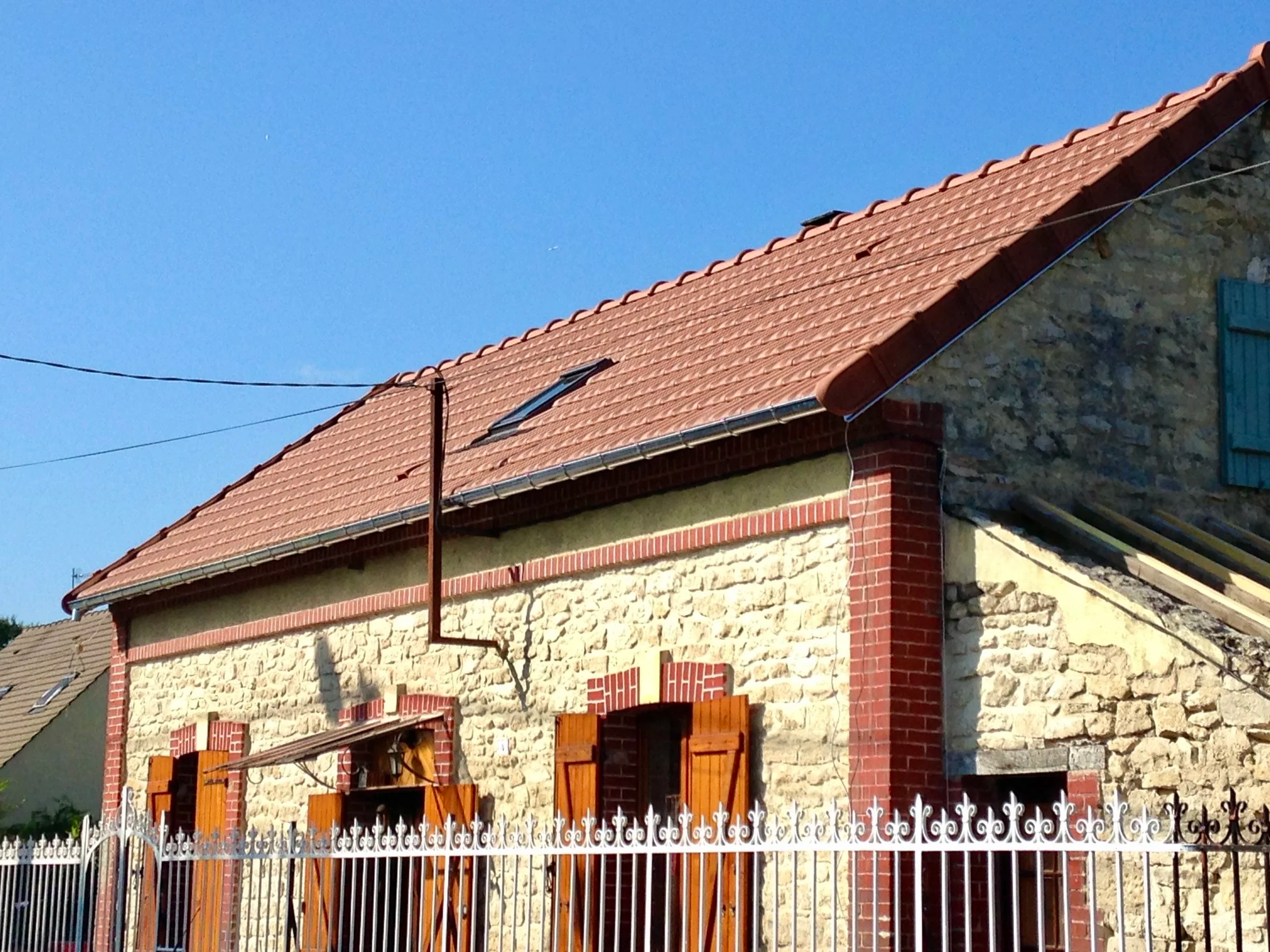 Une maison en pierre avec un toit en tuiles rouges et des volets en bois marron, entourée d'une clôture en métal blanc. Le ciel est clair et bleu.