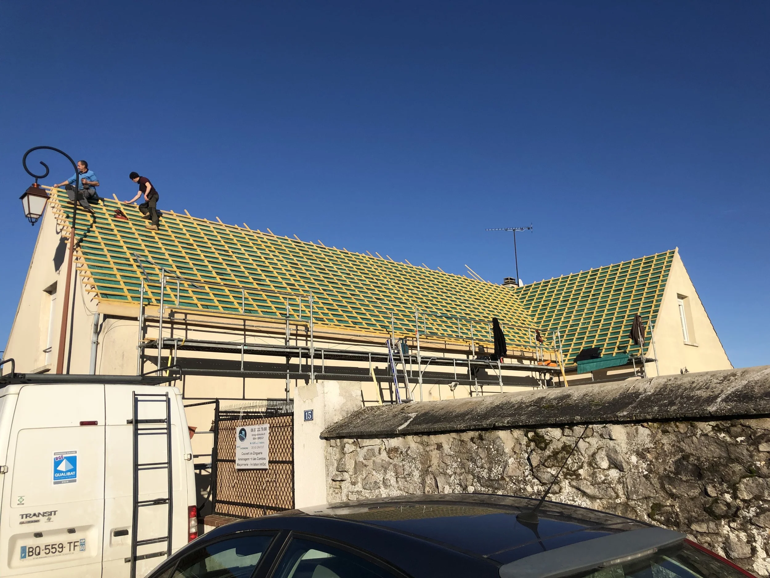 Deux ouvriers travaillent sur la toiture d'une maison en construction, la toiture est recouverte d'une structure en bois, le ciel est dégagé et bleu.