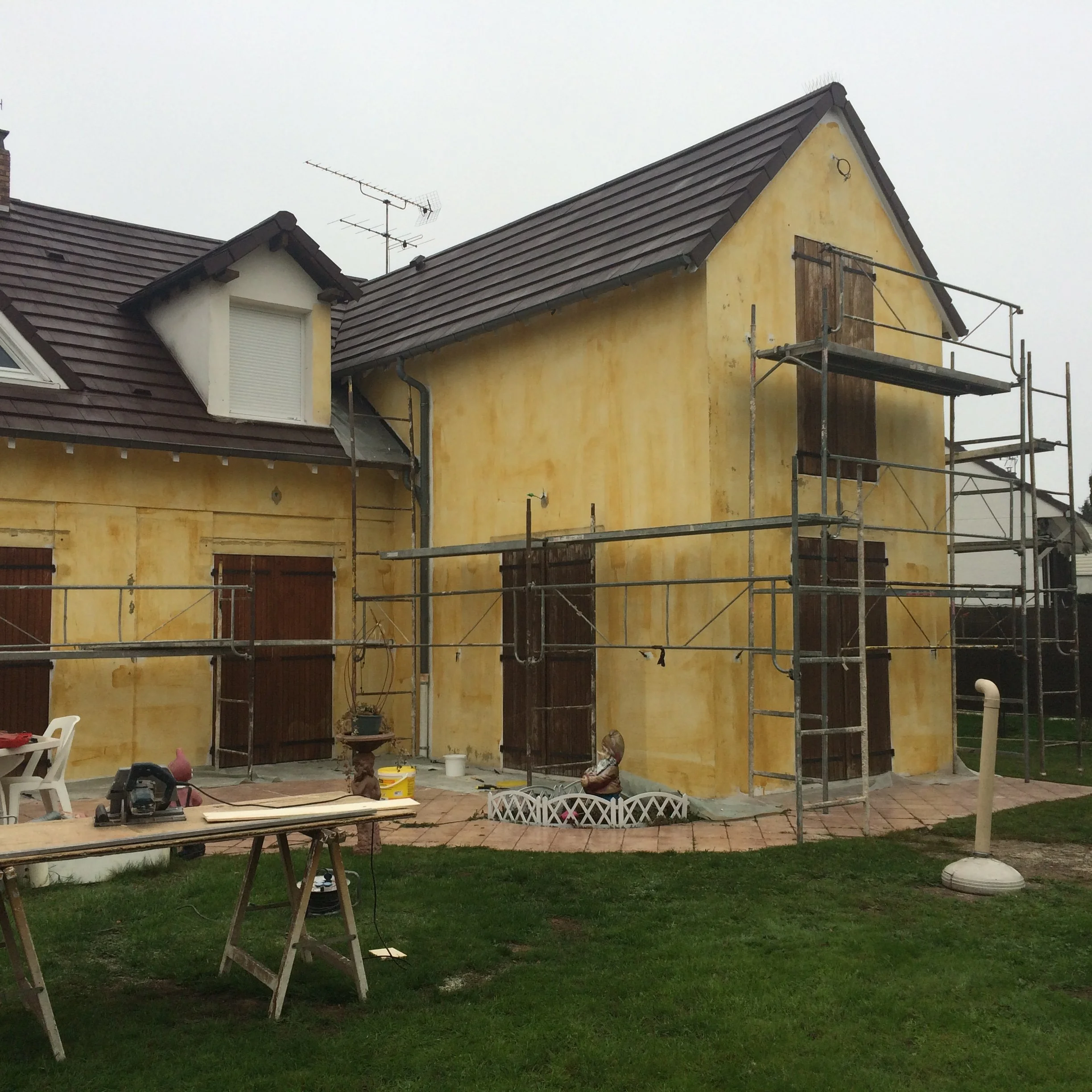 Une maison en construction avec une échafaudage à l'avant, des outils de bricolage sur une table, et un jardin en arrière-plan avec de l'herbe verte.