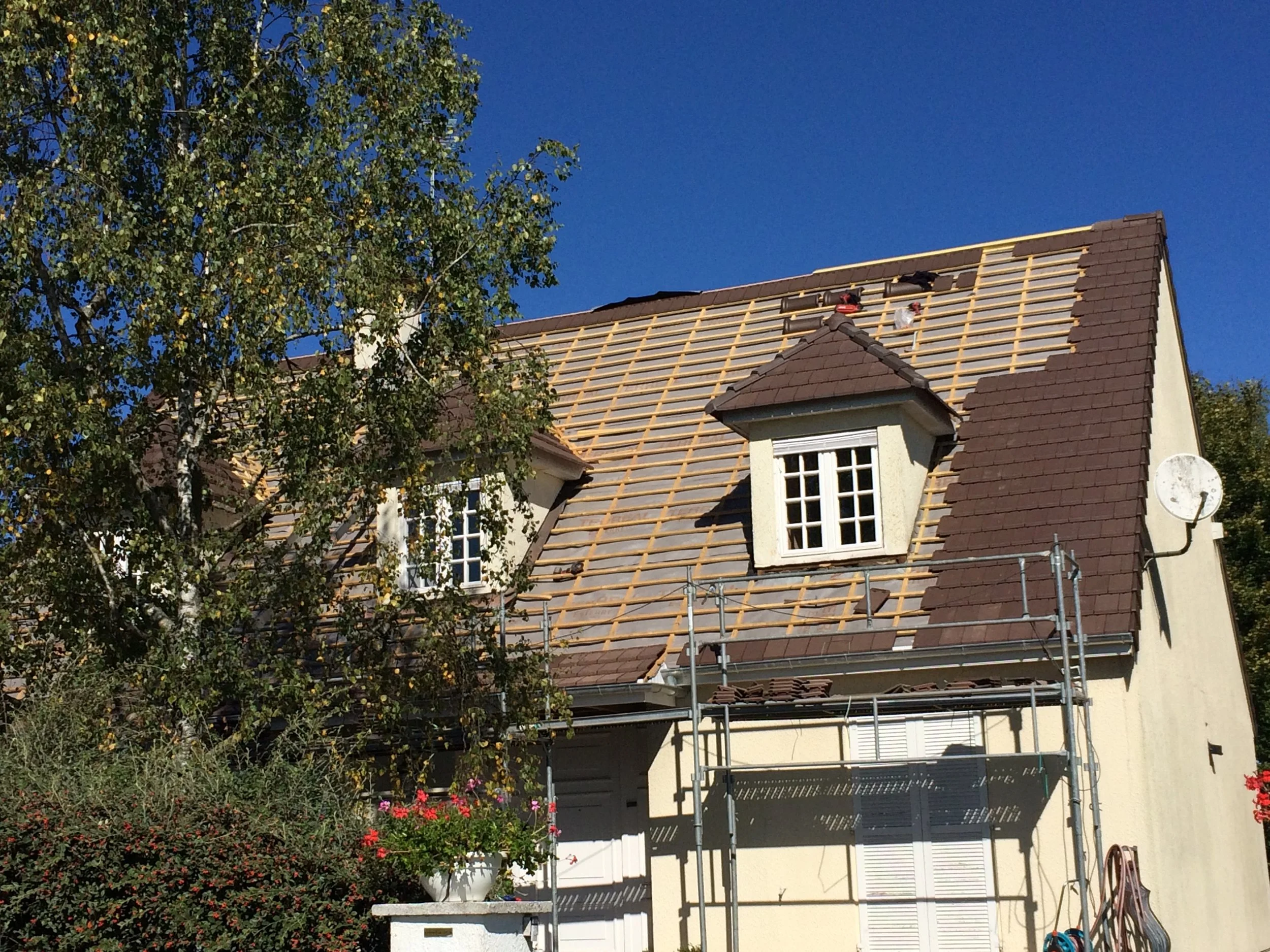 Une maison avec couverture en cours de rénovation, entourée d'arbres et de végétation, sous un ciel bleu clair.