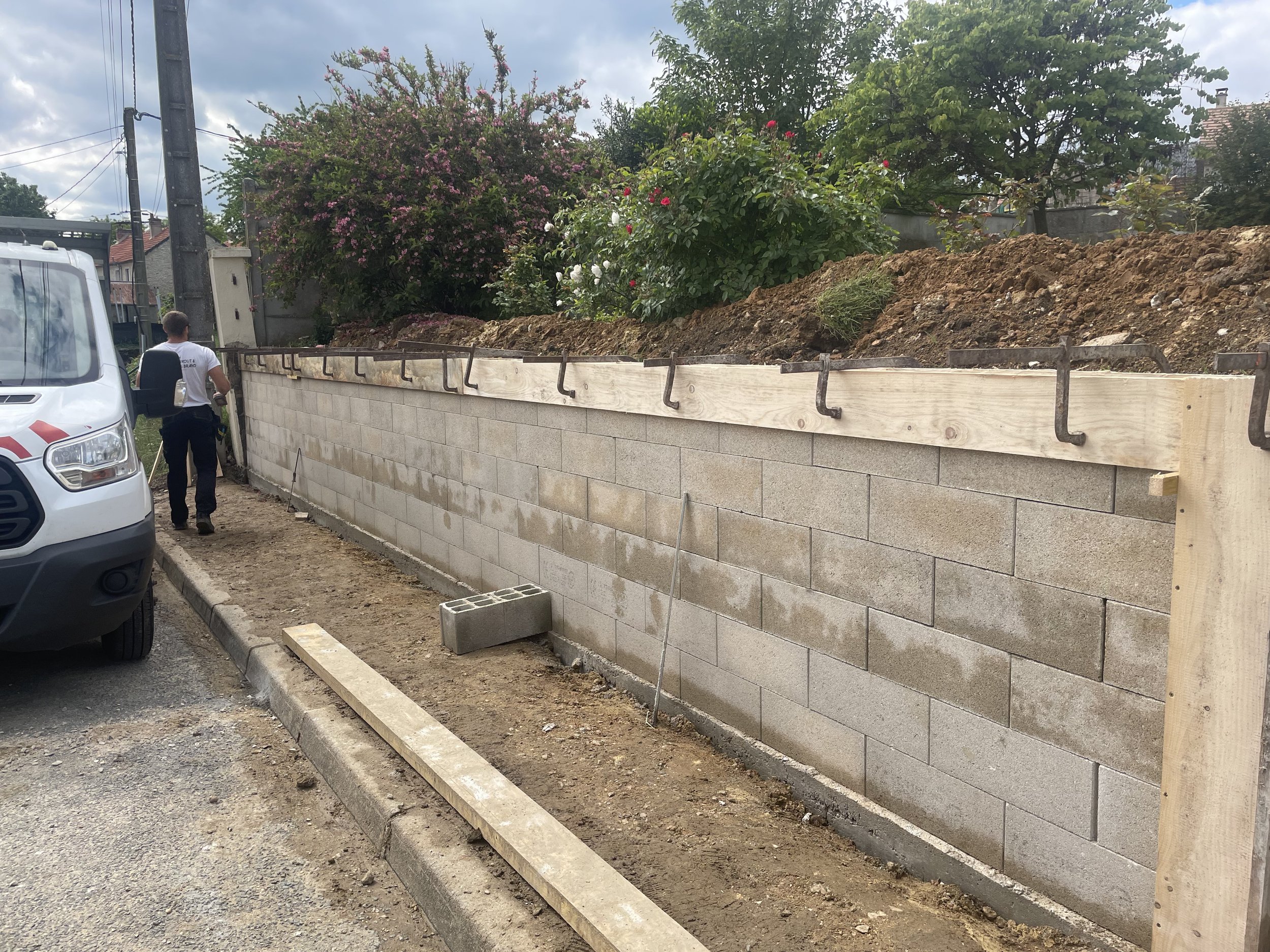 Construction d'un mur en blocs de béton avec un coffrage en bois, en plein jour, dans une zone résidentielle avec des jardins et des arbres en arrière-plan.
