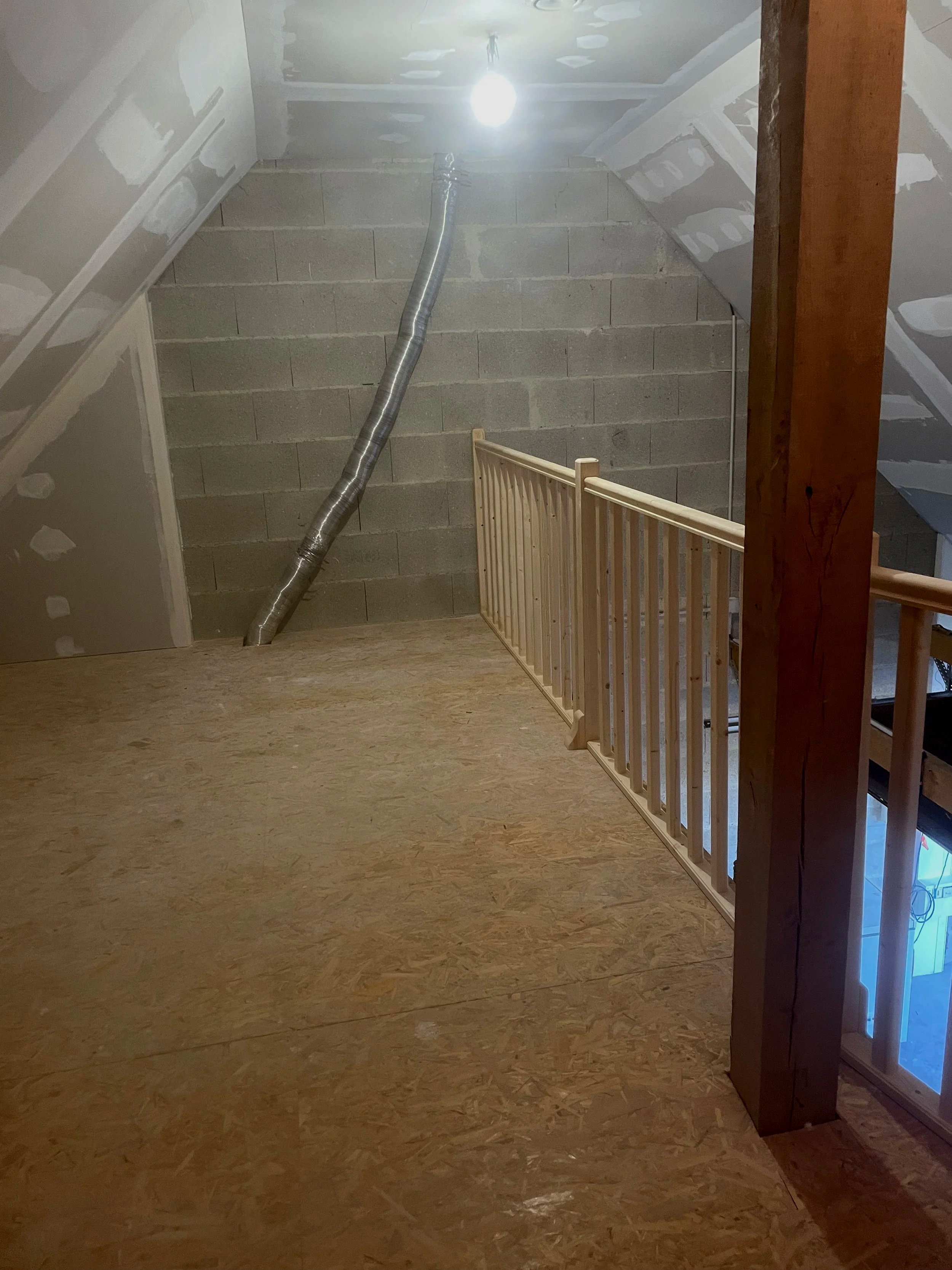 Un espace intérieur en cours de construction avec un mur en bloc de béton, un plafond en plaques de plâtre non fini, un éclairage électrique seul, une rampe en bois, et un tuyau flexible en aluminium qui descend du plafond.