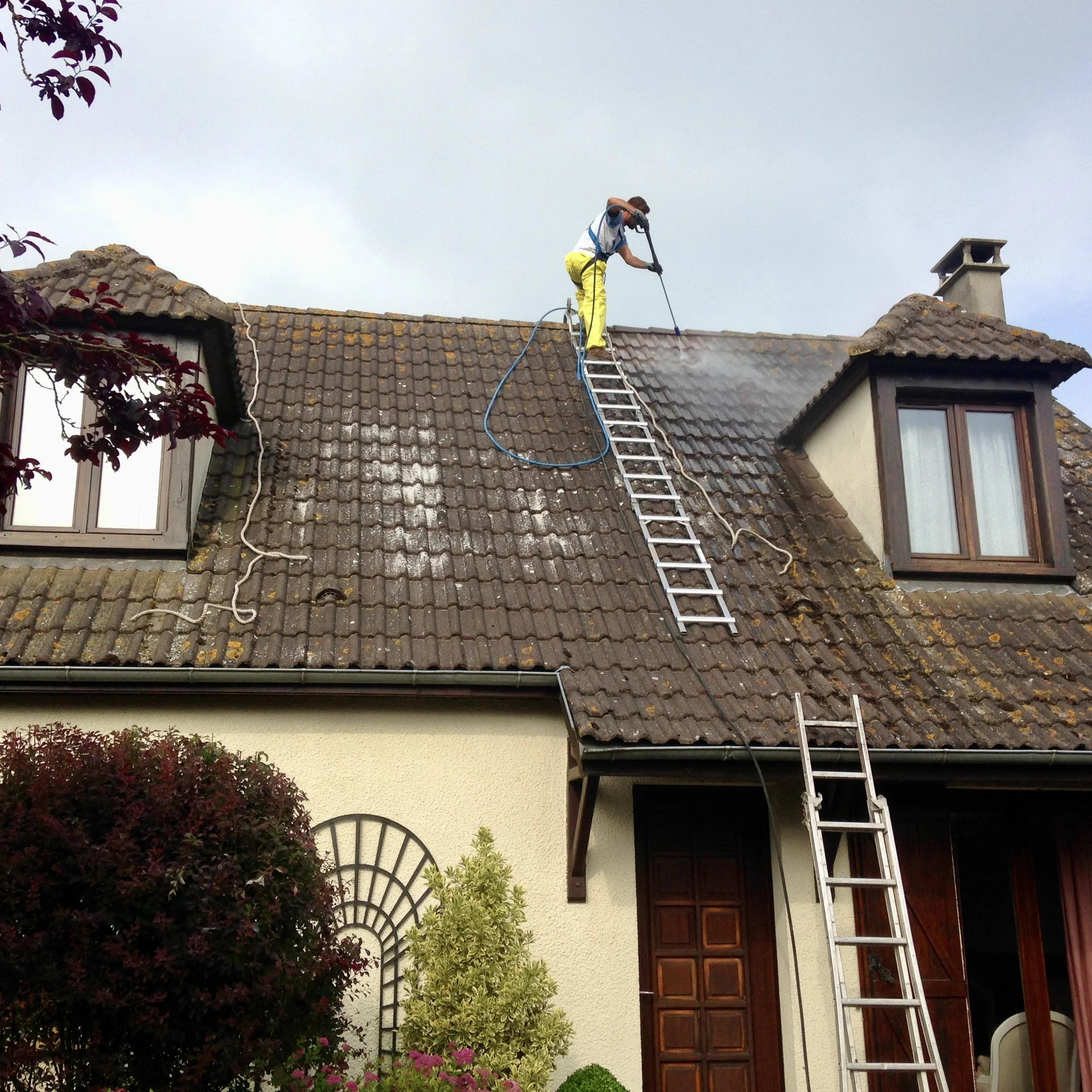 Un homme en vêtements de travail jaune entretien la toiture d'une maison avec un hydrofuge coloré en utilisant une échelle pour accéder au toit.