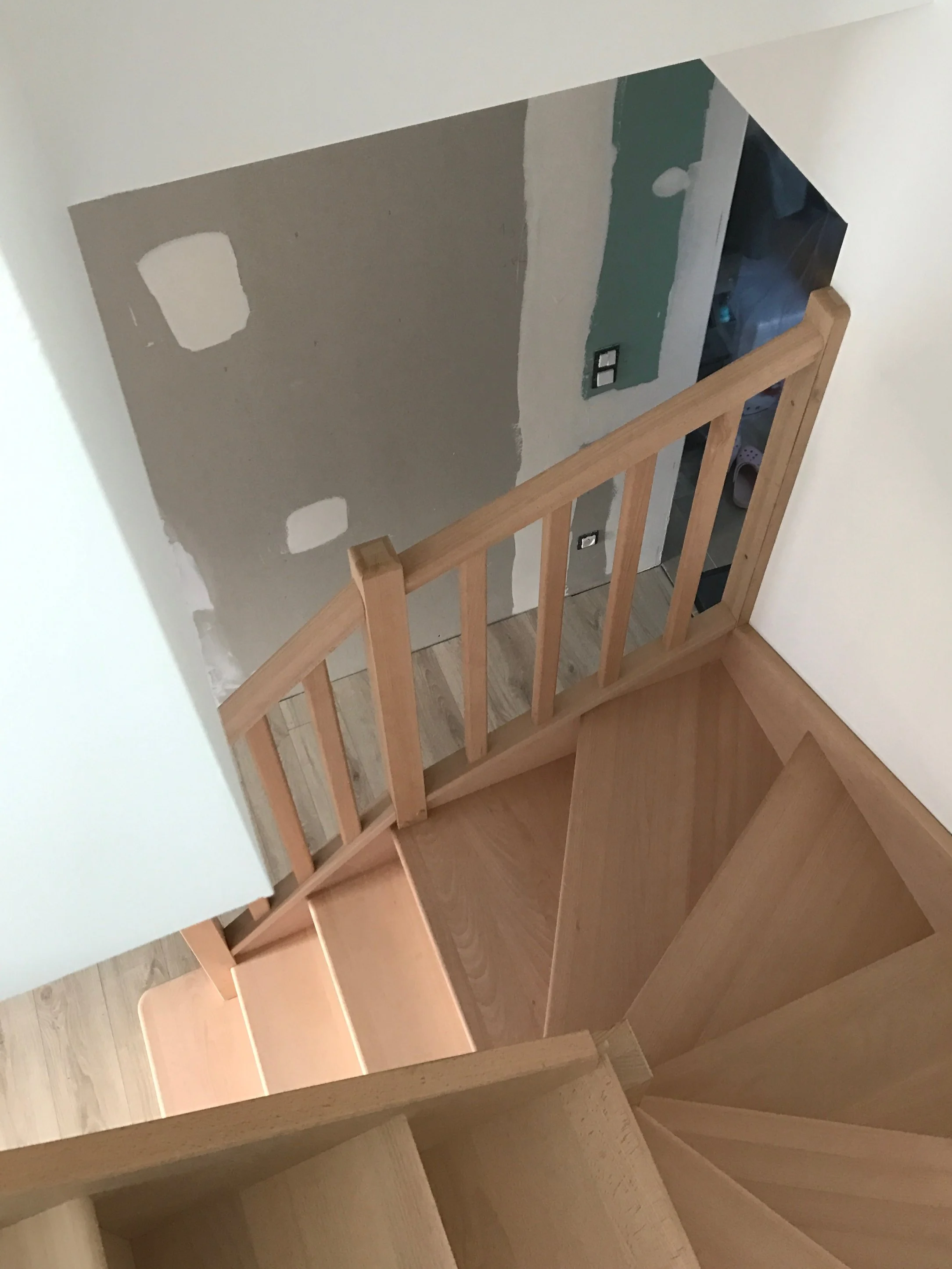 Escalier en bois menant à un étage supérieur avec une rambarde en bois naturel. La photo est prise dans une pièce en cours de rénovation, on voit un mur en plaques de plâtre partiellement peintes ou en cours de préparation, avec deux prises électriques encastrées. L'ambiance est lumineuse avec un sol en parquet.