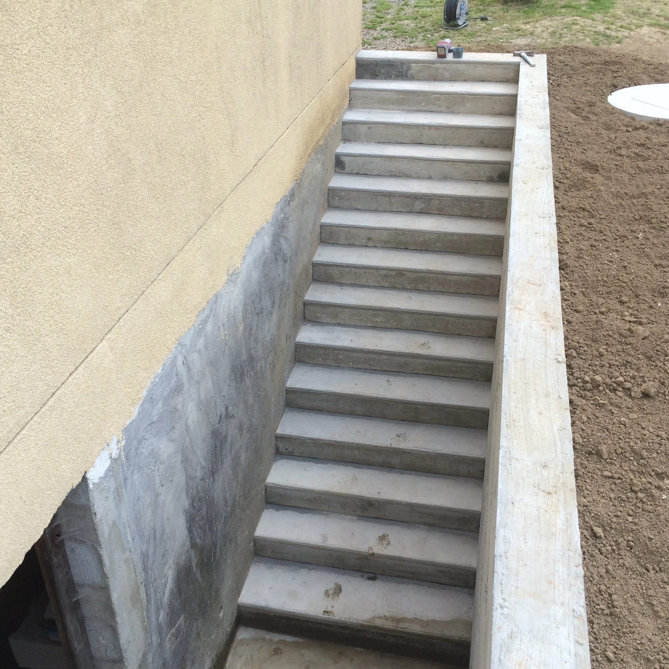 Escalier en béton en cours de construction avec une paroi murale beige à gauche, des outils et matériaux à proximité, et un terrain en terre à droite.