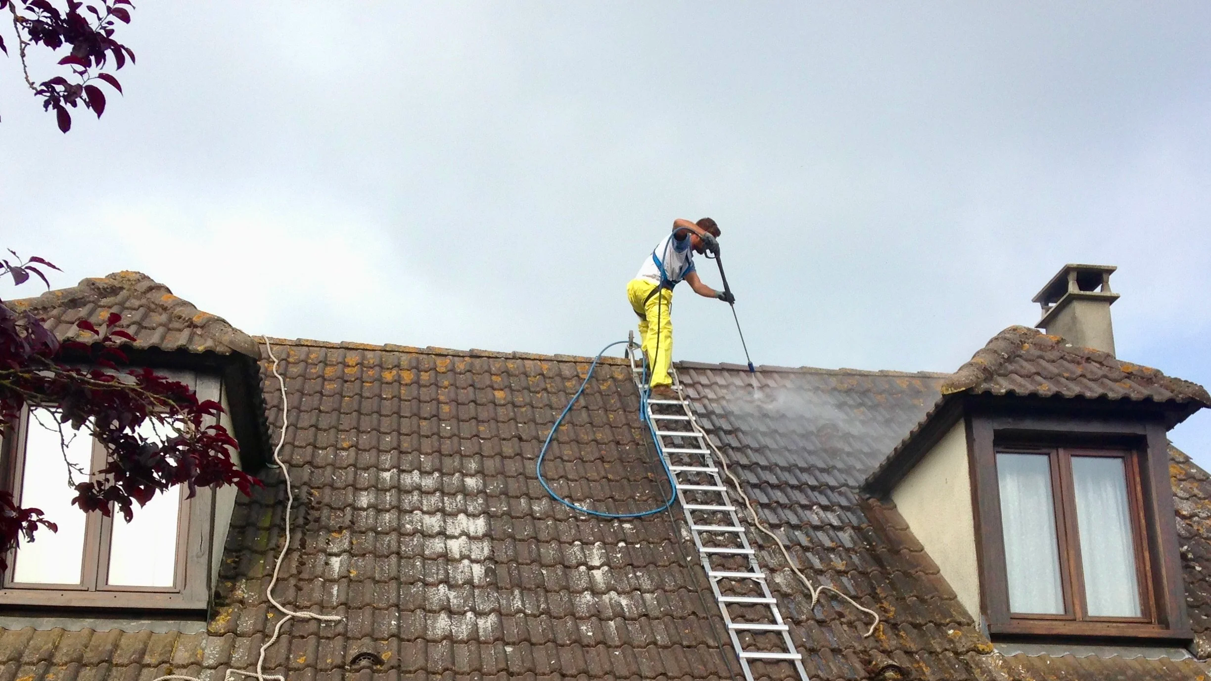 Un homme en vêtements de travail jaune entretien la toiture d'une maison avec un hydrofuge coloré en utilisant une échelle pour accéder au toit.