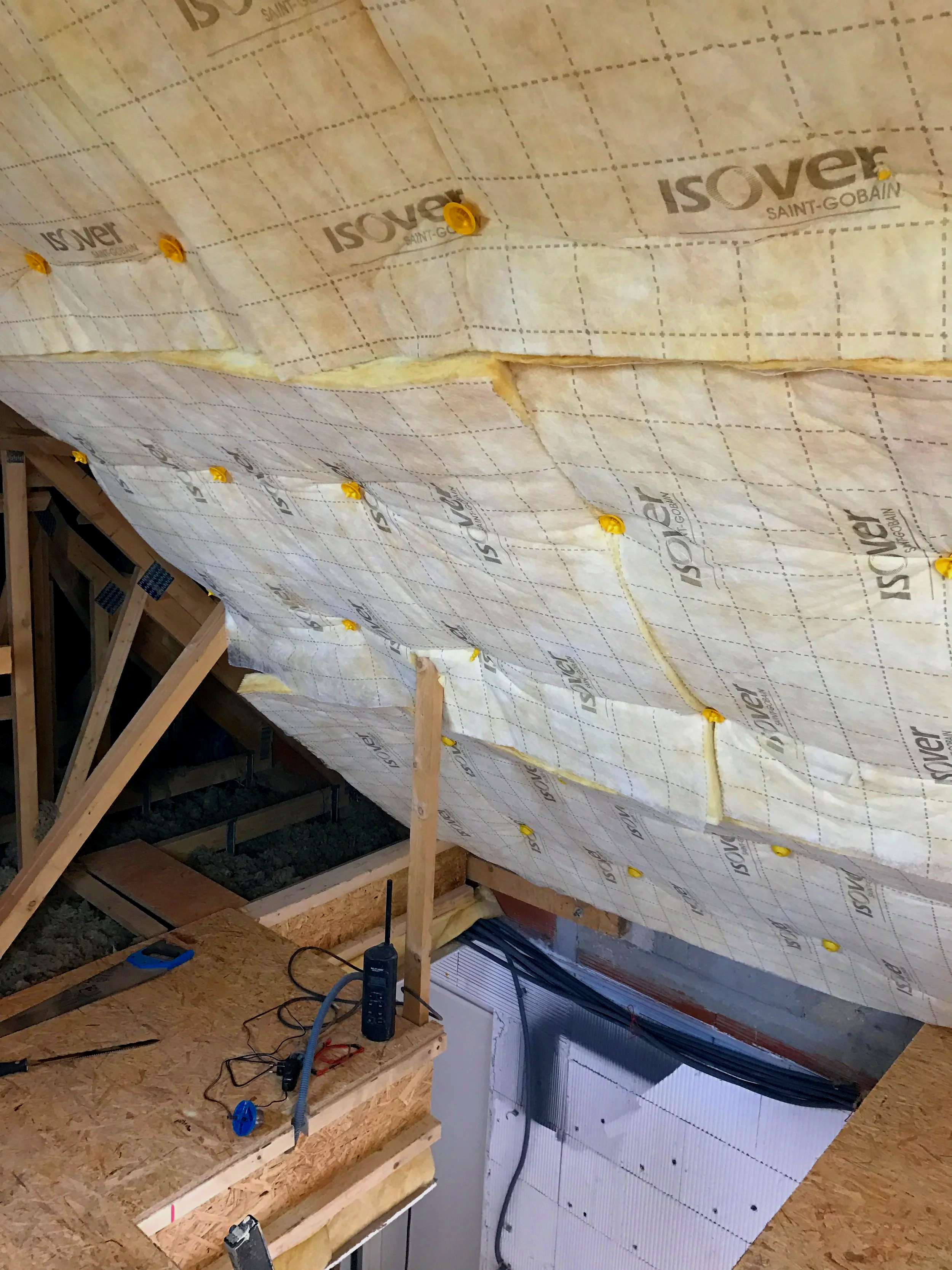 Photo montrant l'intérieur d'une toiture en construction avec une isolation en mousse de polyuréthane et des structures en bois. Des outils et câbles se trouvent sur un support en bois au premier plan.