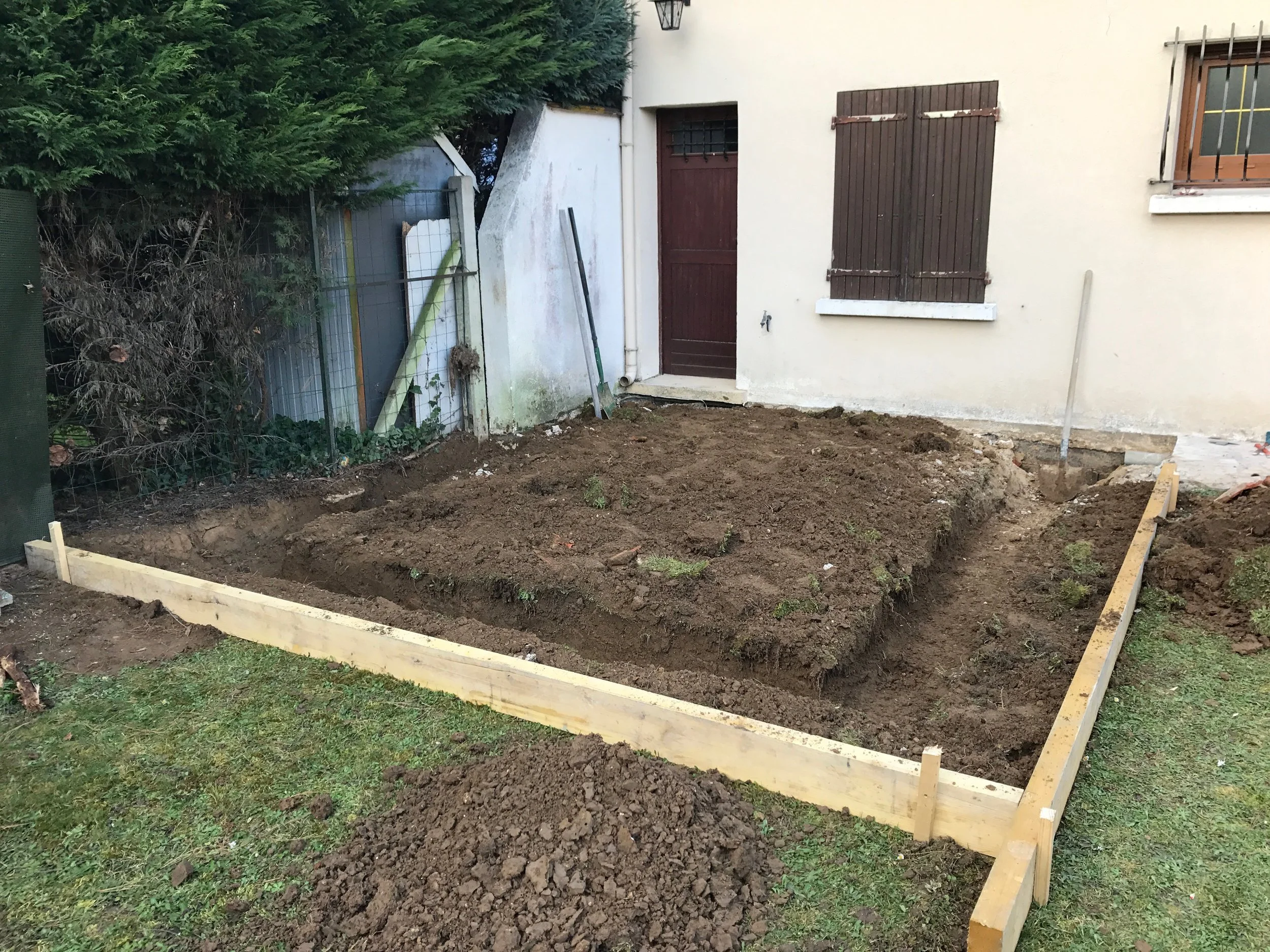 Terrassement d'une zone de jardin entourée d'une clôture en grillage, avec une maison en fond, fenêtres avec volets en bois, et une grande haie d'arbres. Le sol est préparé avec des contours en bois pour une nouvelle construction ou aménagement.