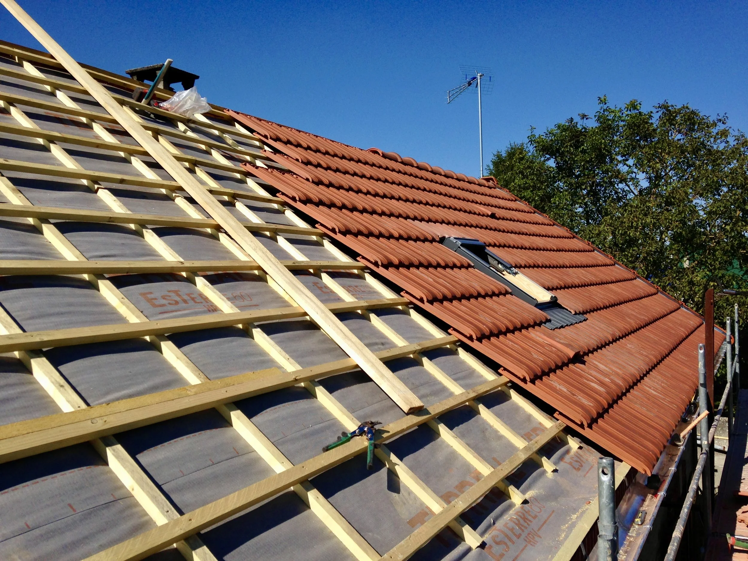 Toit en cours de rénovation avec des tuiles rouges partiellement installées et un litonnage pour supporter les futures tuiles, sous un ciel clair et un arbre vert à côté.