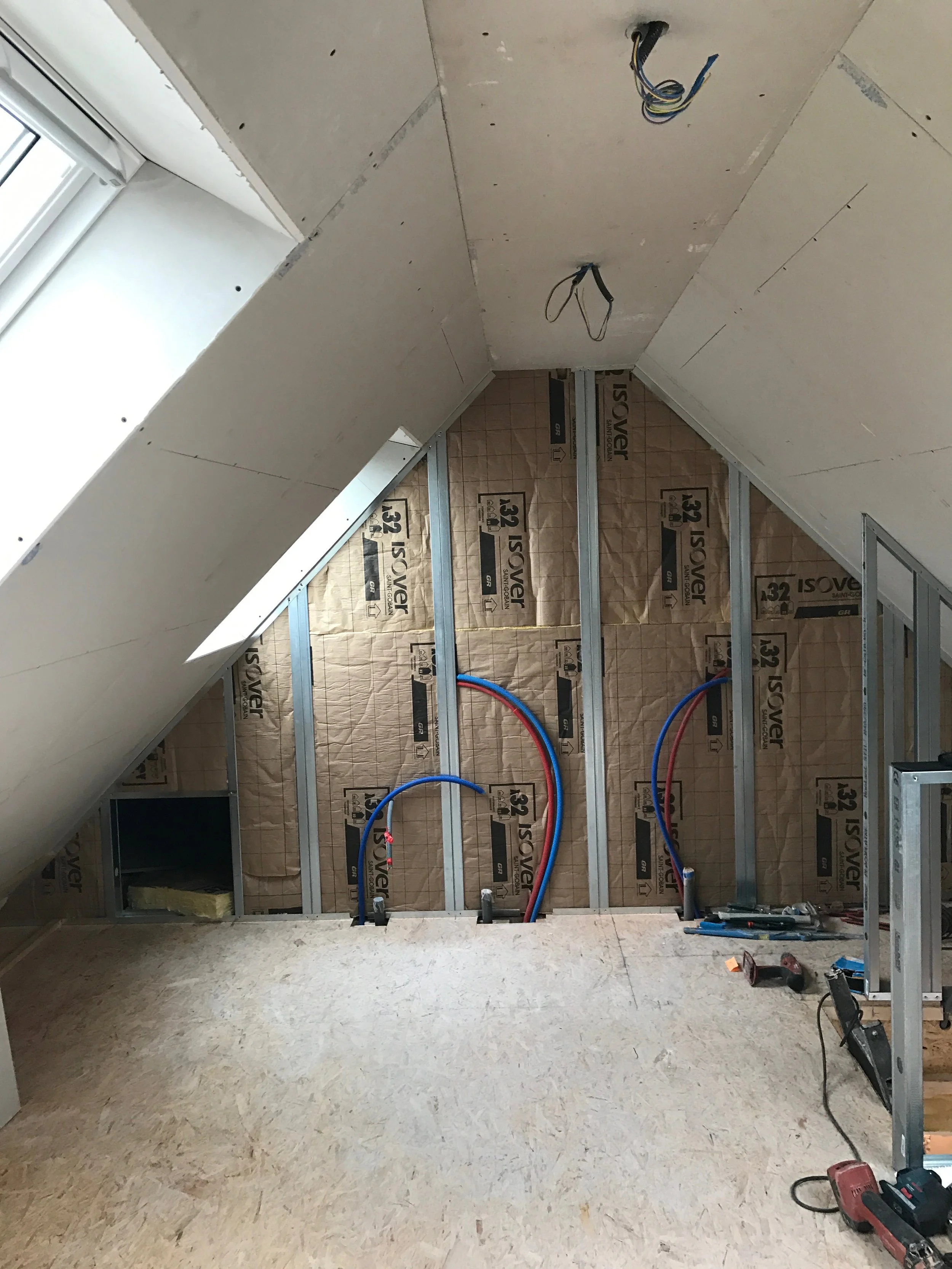 Salle en cours de construction avec murs en plaques de plâtre et installation électrique en cours, tuyaux rouges et bleus visibles, outils et matériaux divers présents au sol.
