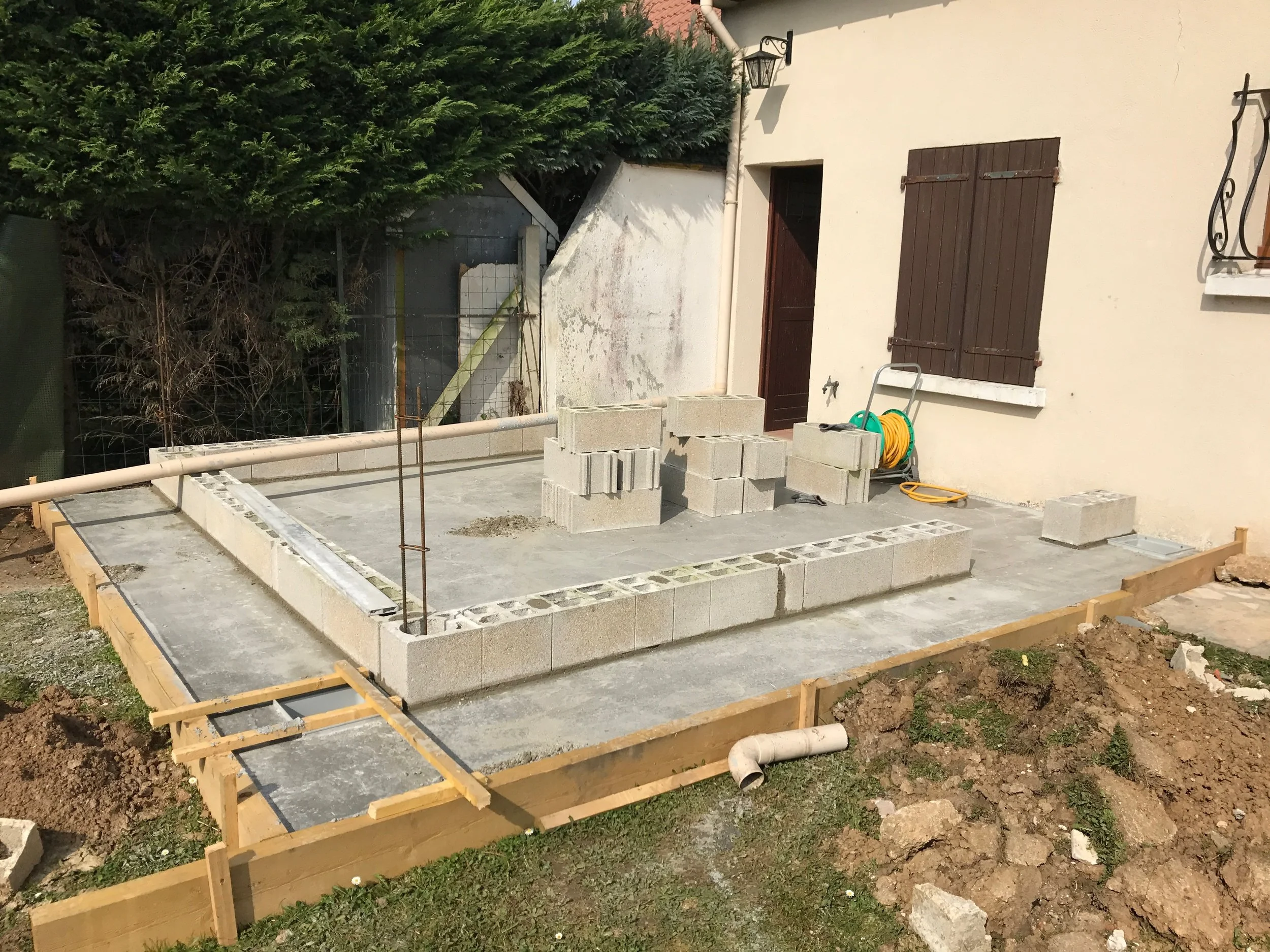 Construction d'une nouvelle terrasse avec dalle et blocs de béton, un coffrage en bois, des outils et un tuyau d'arrosage, près d'une maison avec volets en bois.