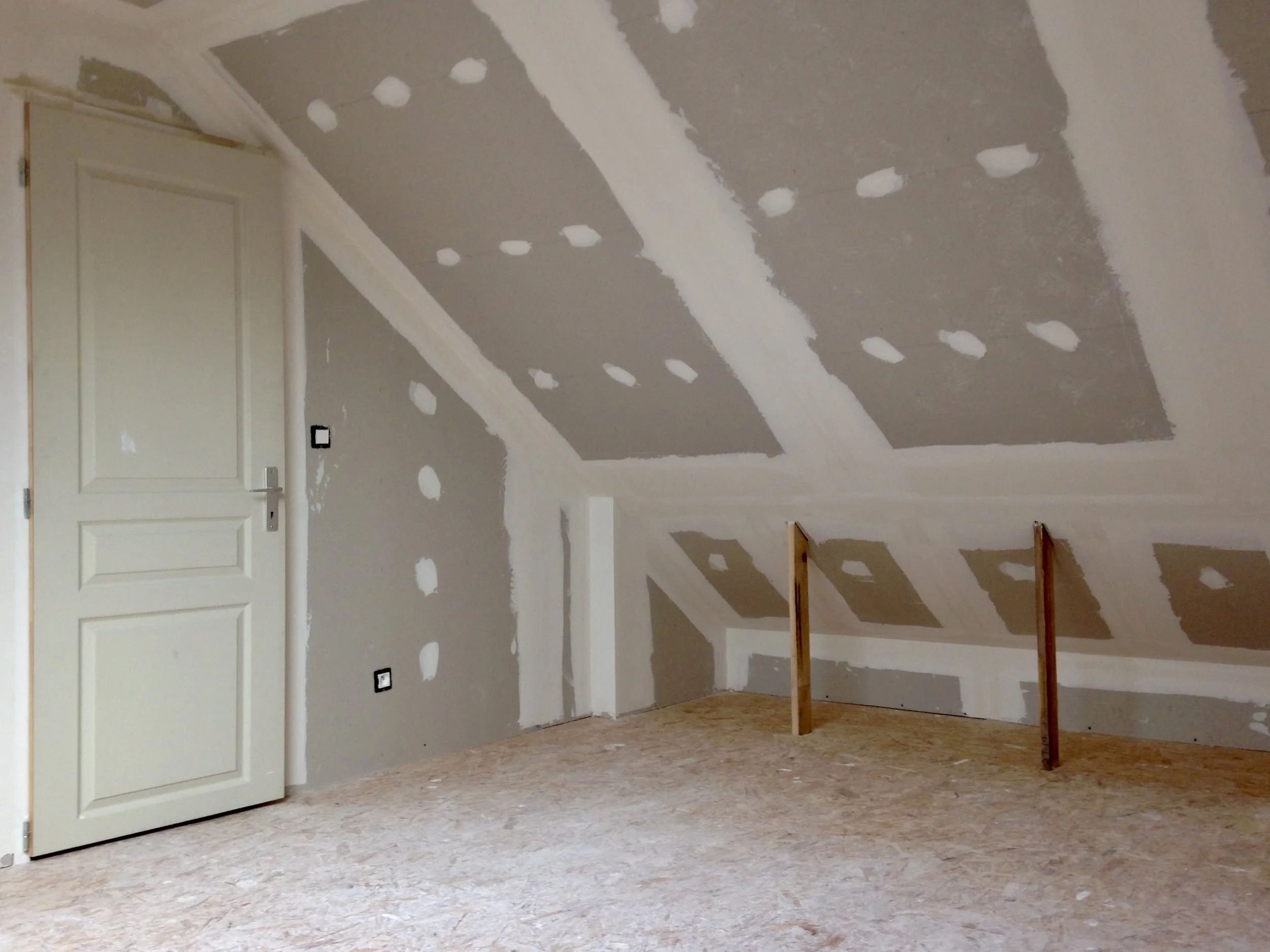 Une pièce sous construction avec des murs en plaque de plâtre et une cloison en ossature bois, le sol est en panneaux de particules.
