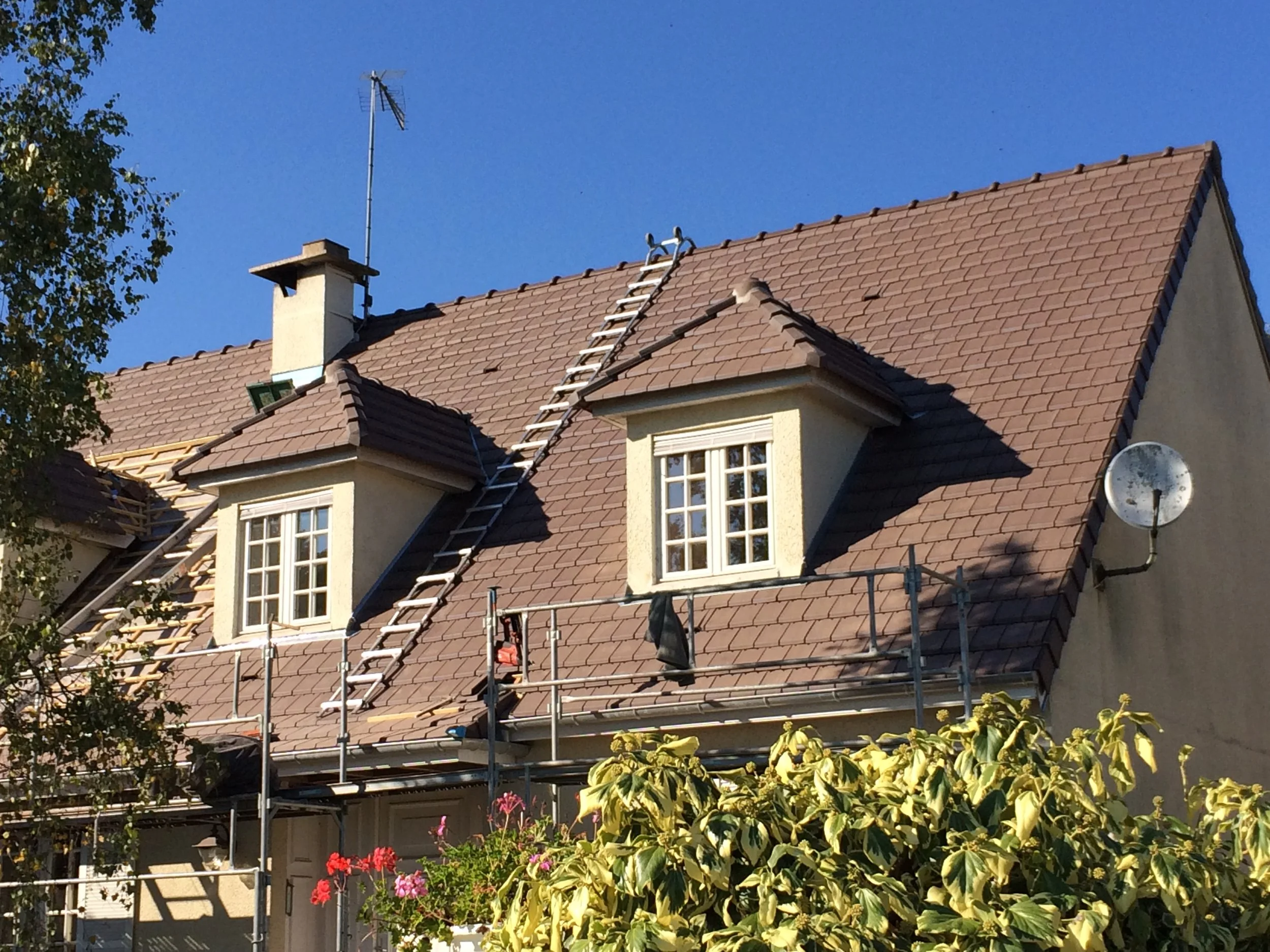 Une maison avec une nouvelle couverture, des fenêtres blanches et un échafaudage. Sur le toit, un échelle est appuyée contre la pente. Le ciel est bleu clair sans nuages, et il y a des arbres et des fleurs en premier plan.