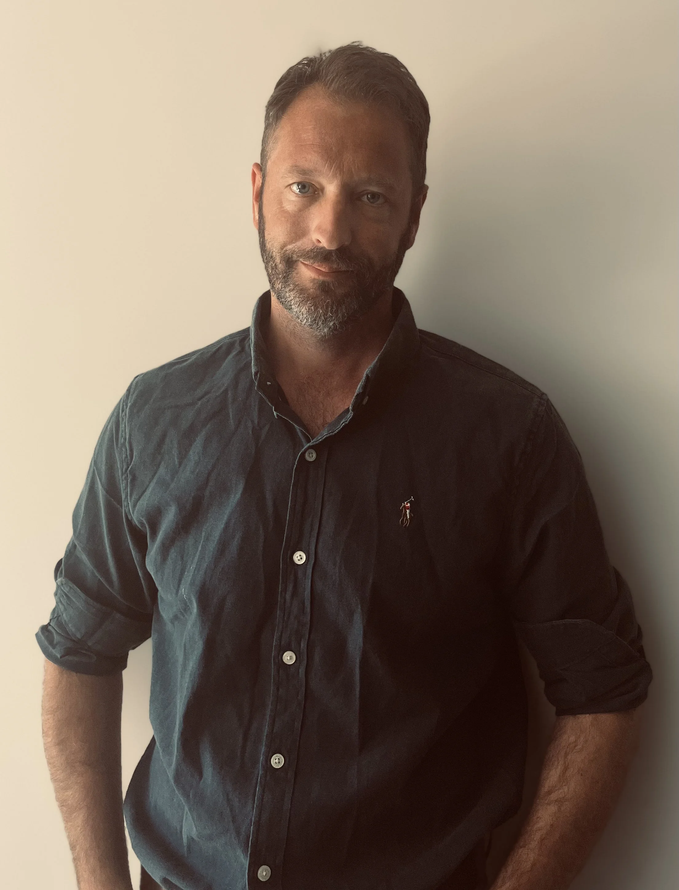 A man with dark hair and a beard, wearing a dark button-up shirtsleeve, standing against a plain beige wall.
