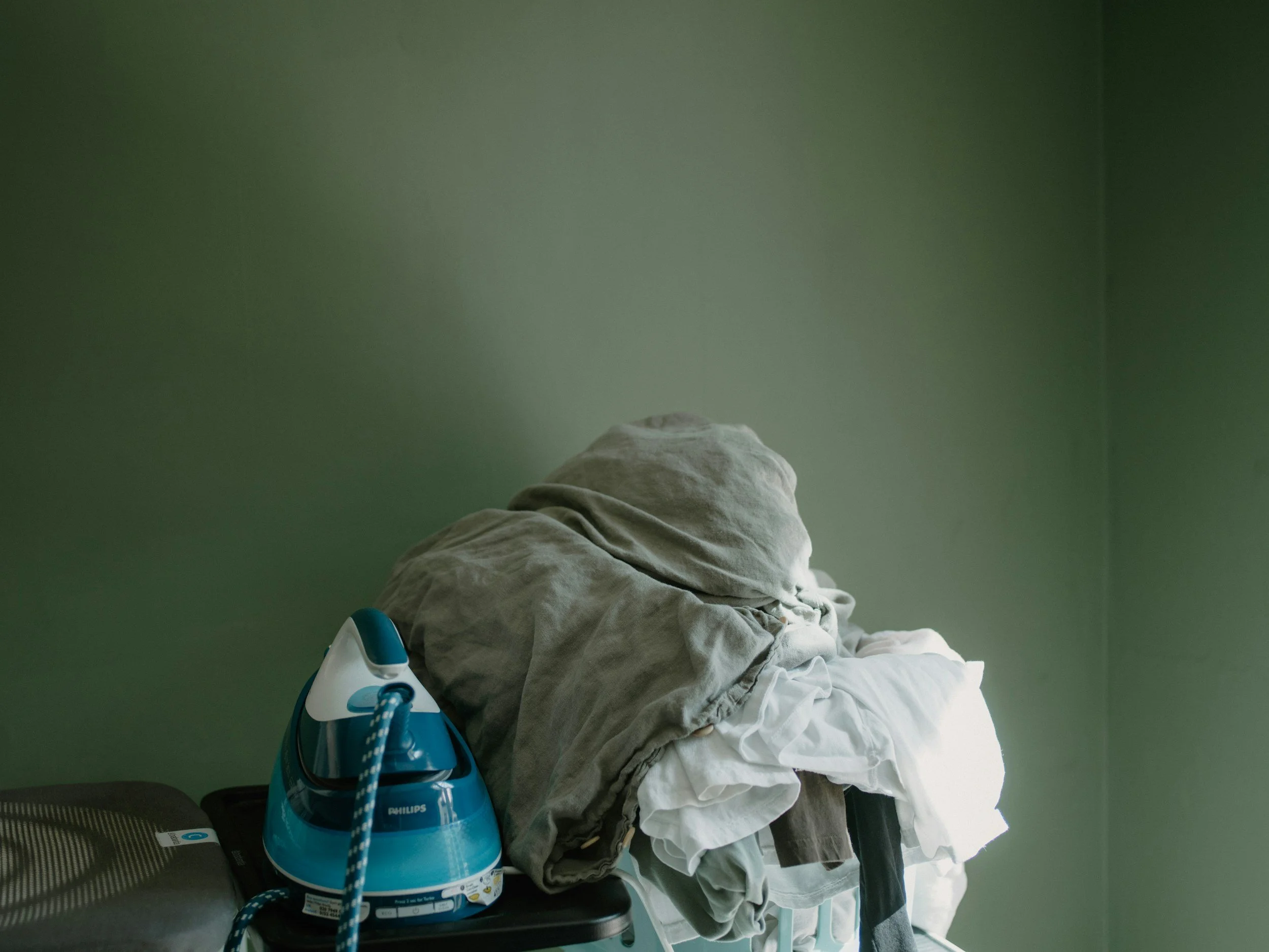 A bed with a pile of beige and white blankets against a green wall, and a blue steam iron resting on the bed.