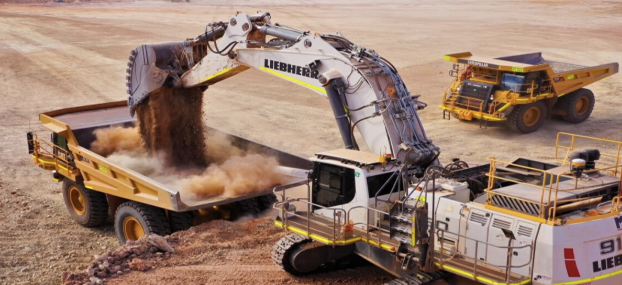 An excavator with a Liebherr logo is dumping dirt into a dump truck, with a large yellow mining truck nearby, in a desert terrain.