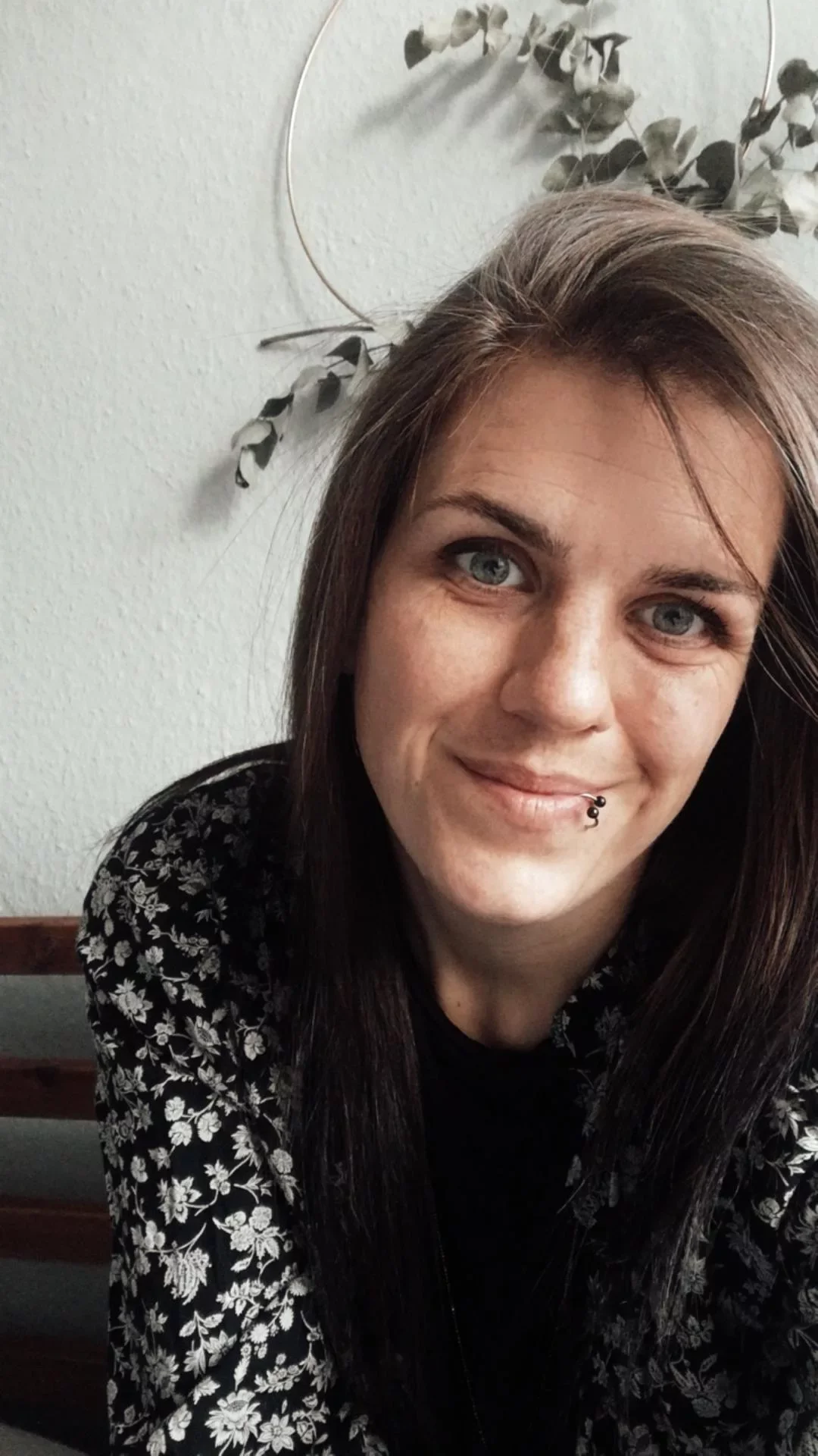 Eine lächelnde Frau mit Piercing in der Unterlippe, dunkelbraune Haare und eine schwarze Bluse mit weißen Blumen, sitzend auf einer Bank vor weißer Wand mit dekorativem Wandhänger.