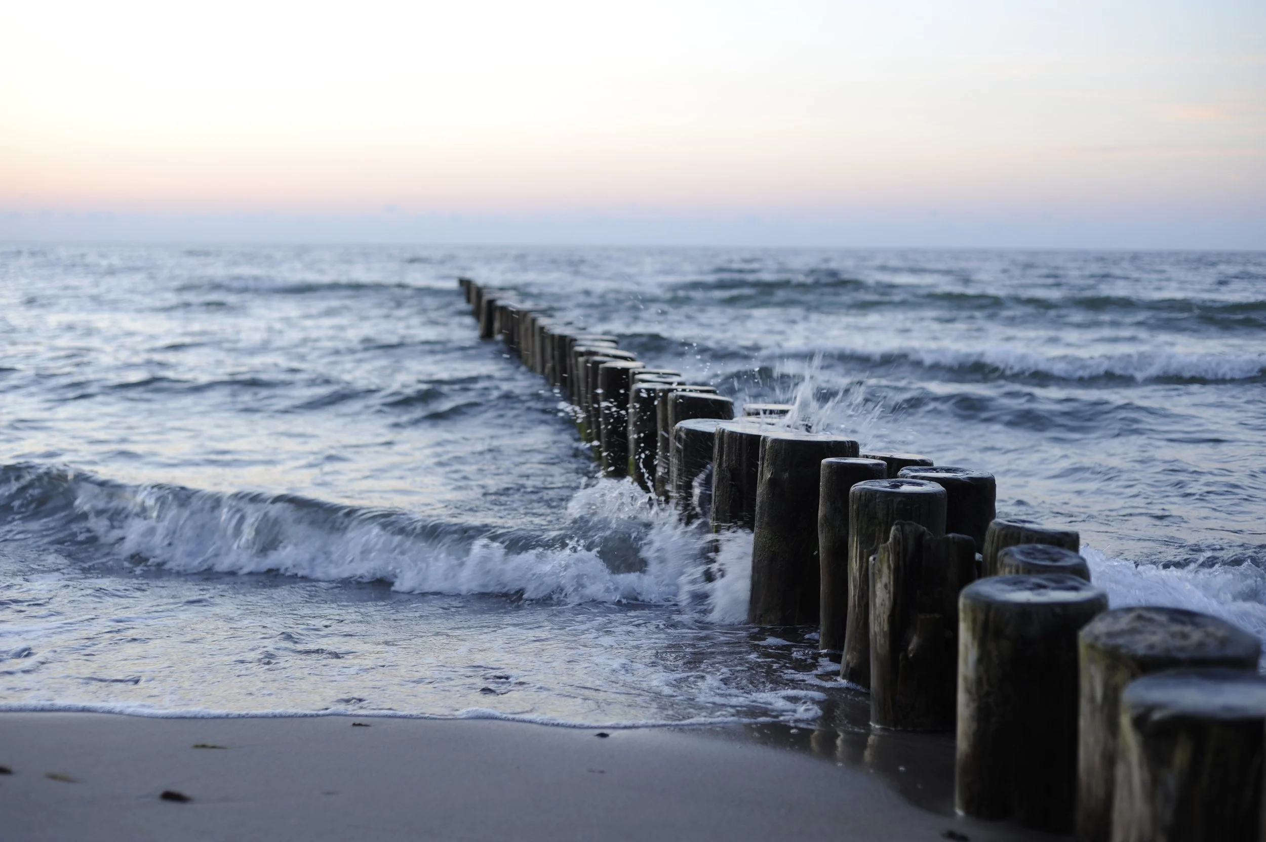 Ein Meer mit Wellen und ein Himmel bei Sonnenuntergang, im Vordergrund eine Reihe von Holzpfählen im Wasser.