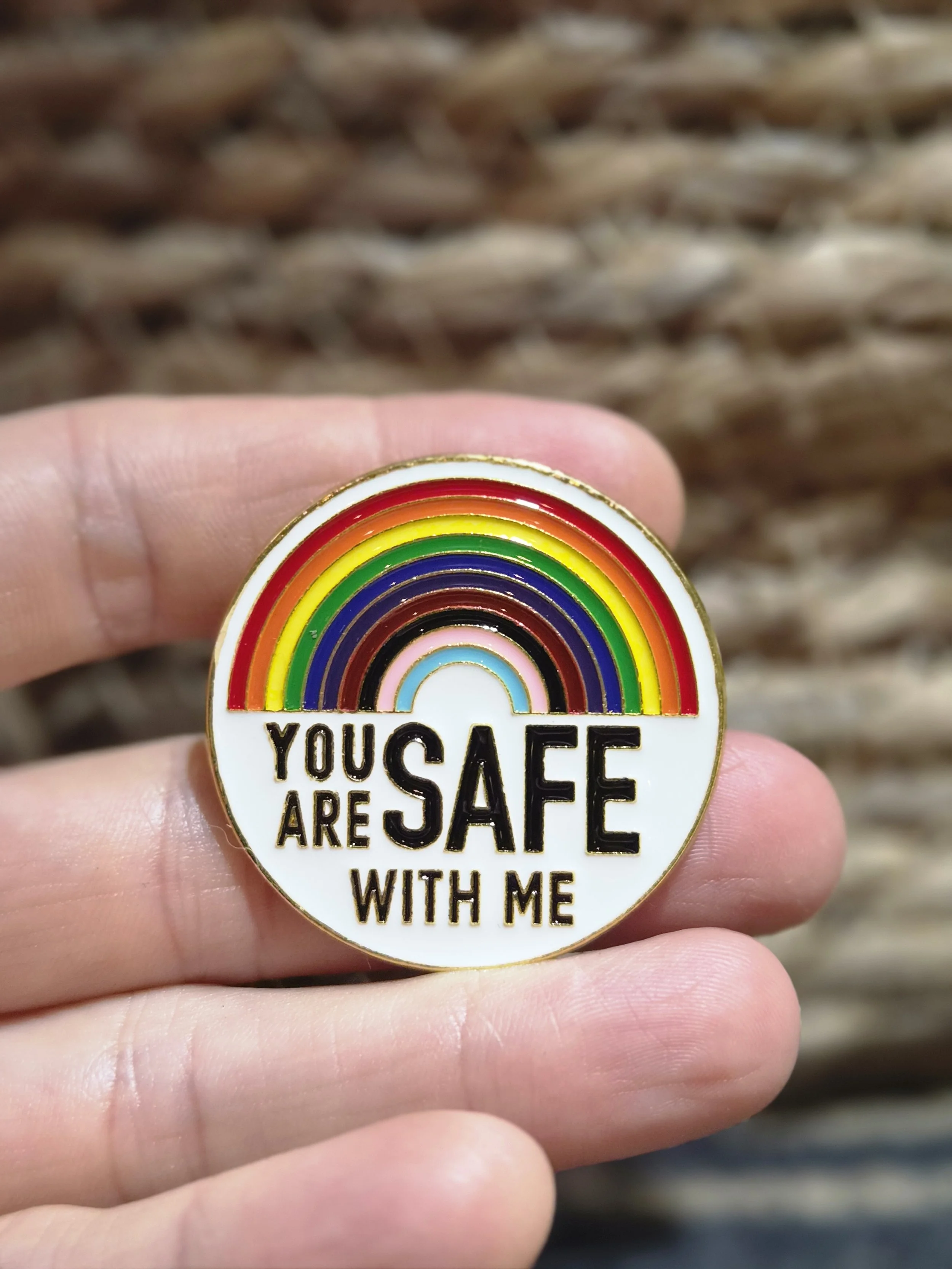 Eine Hand hält einen runden Pin mit einem Regenbogenmotiv und dem Schriftzug 'You Are Safe With Me' vor einem braunen, geflochtenen Hintergrund.