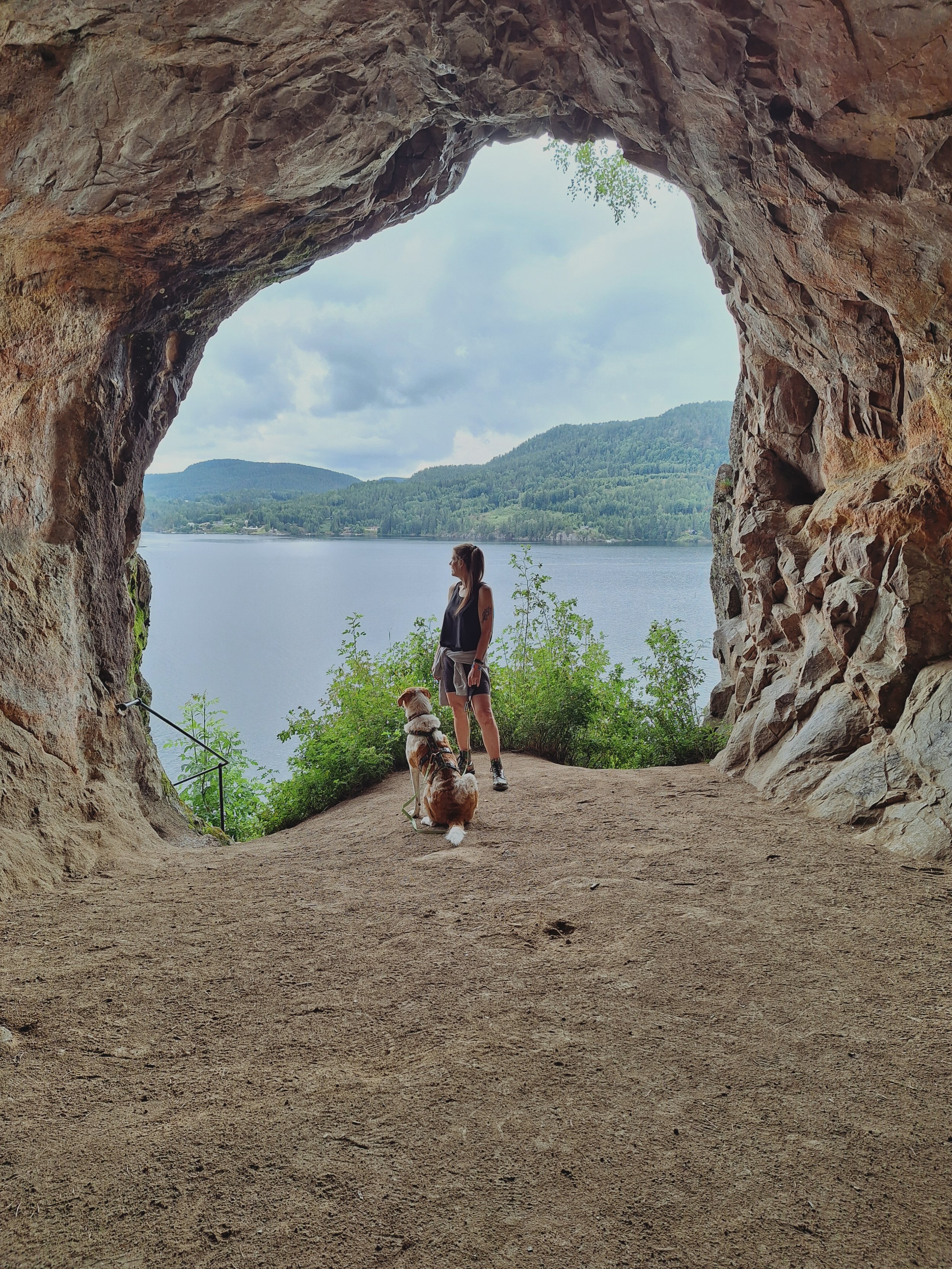 Frau mit Hund vor einem Wassersee, sichtbar durch eine Felshöhle mit Blick auf die Landschaft.