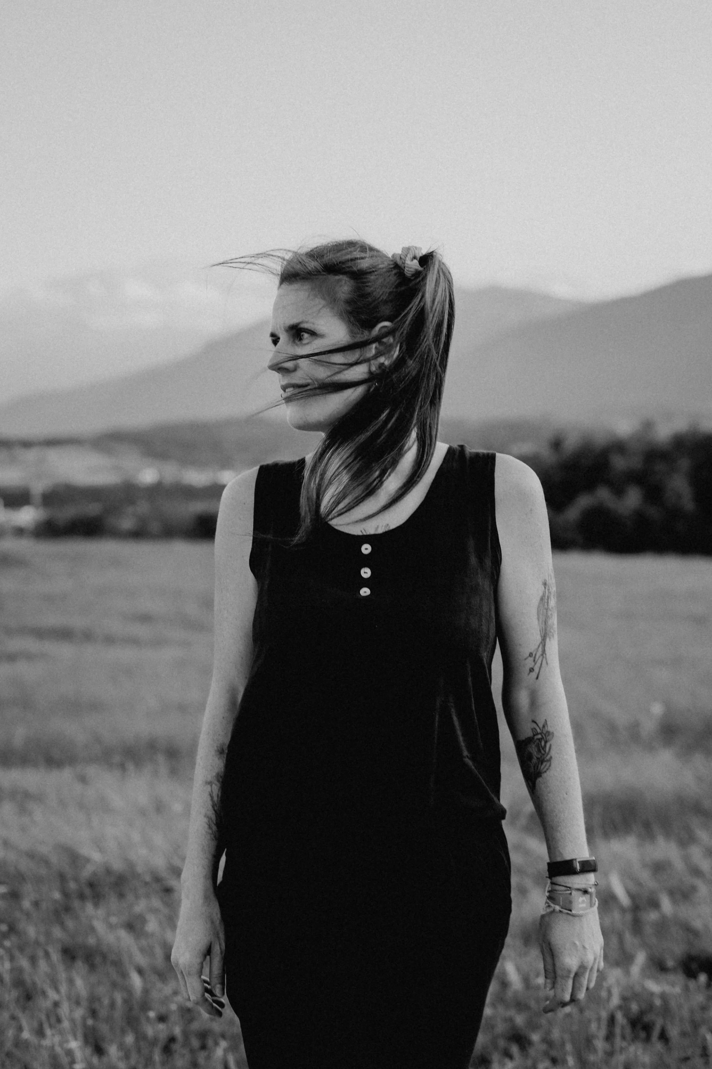 Schwarz-Weiß-Foto einer jungen Frau mit langem Haar in einem freien Feld, die nach rechts schaut, bei windigem Wetter, mit Tätowierungen auf den Armen, trägt ein schwarzes Shirt und Armbänder.