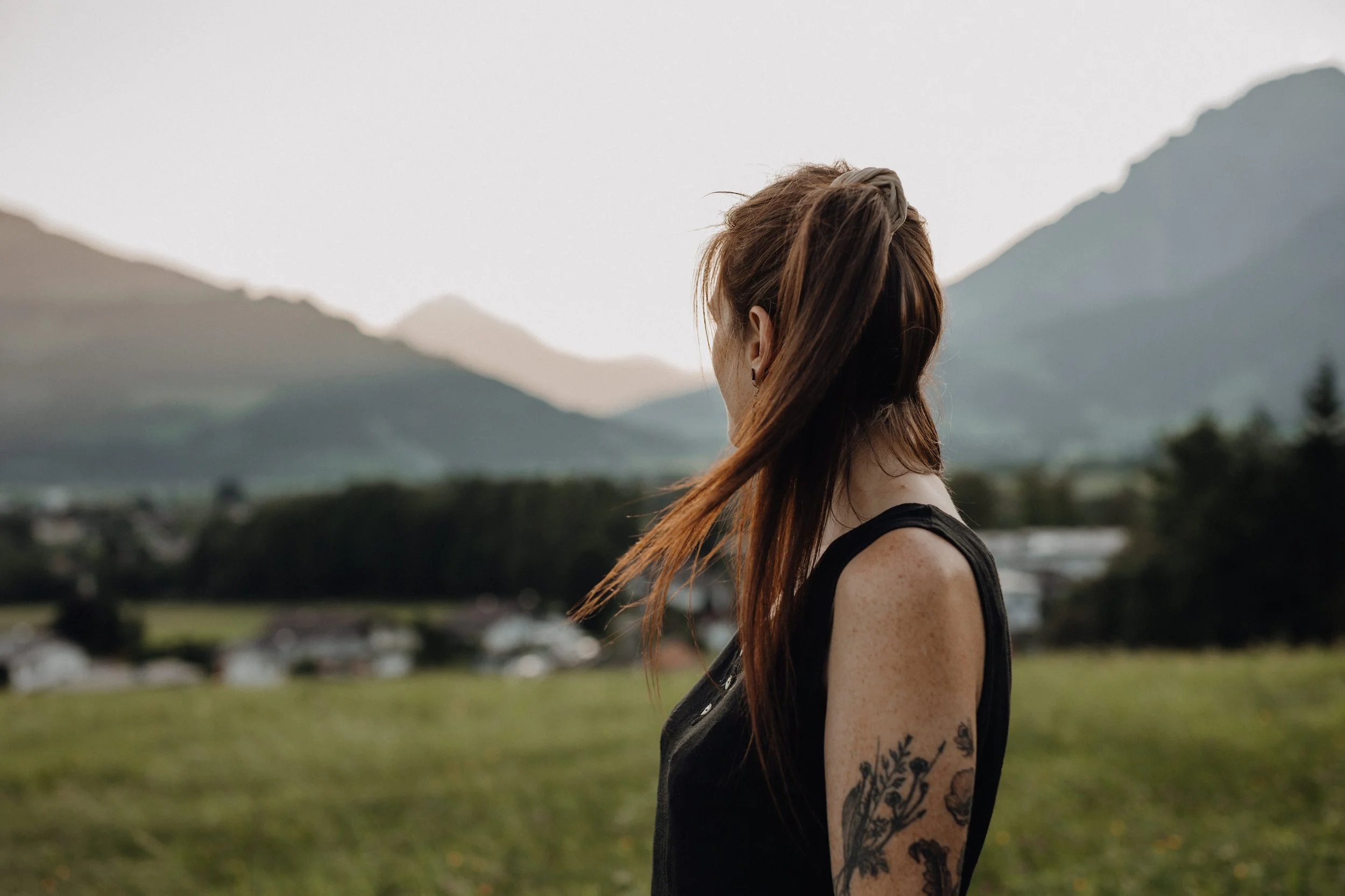 Seitenansicht einer jungen Frau mit Tattoos am Arm auf einer grünen Wiese, Berge im Hintergrund, bewölkter Himmel.