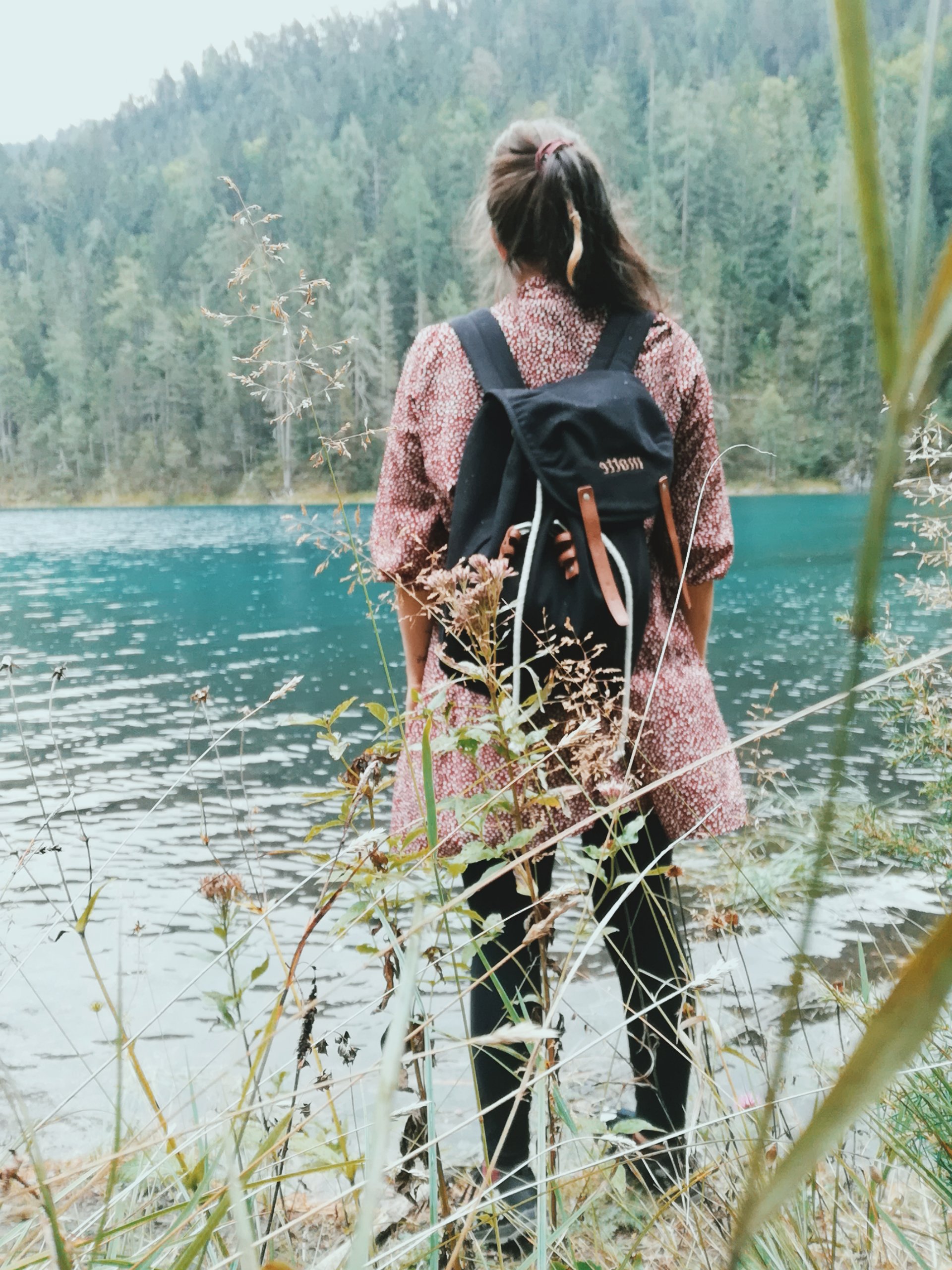 Eine Frau mit braunem, lockigem Haar steht an einem See, umgeben von Bäumen, und trägt einen schwarzen Rucksack. Sie schaut auf das Wasser.