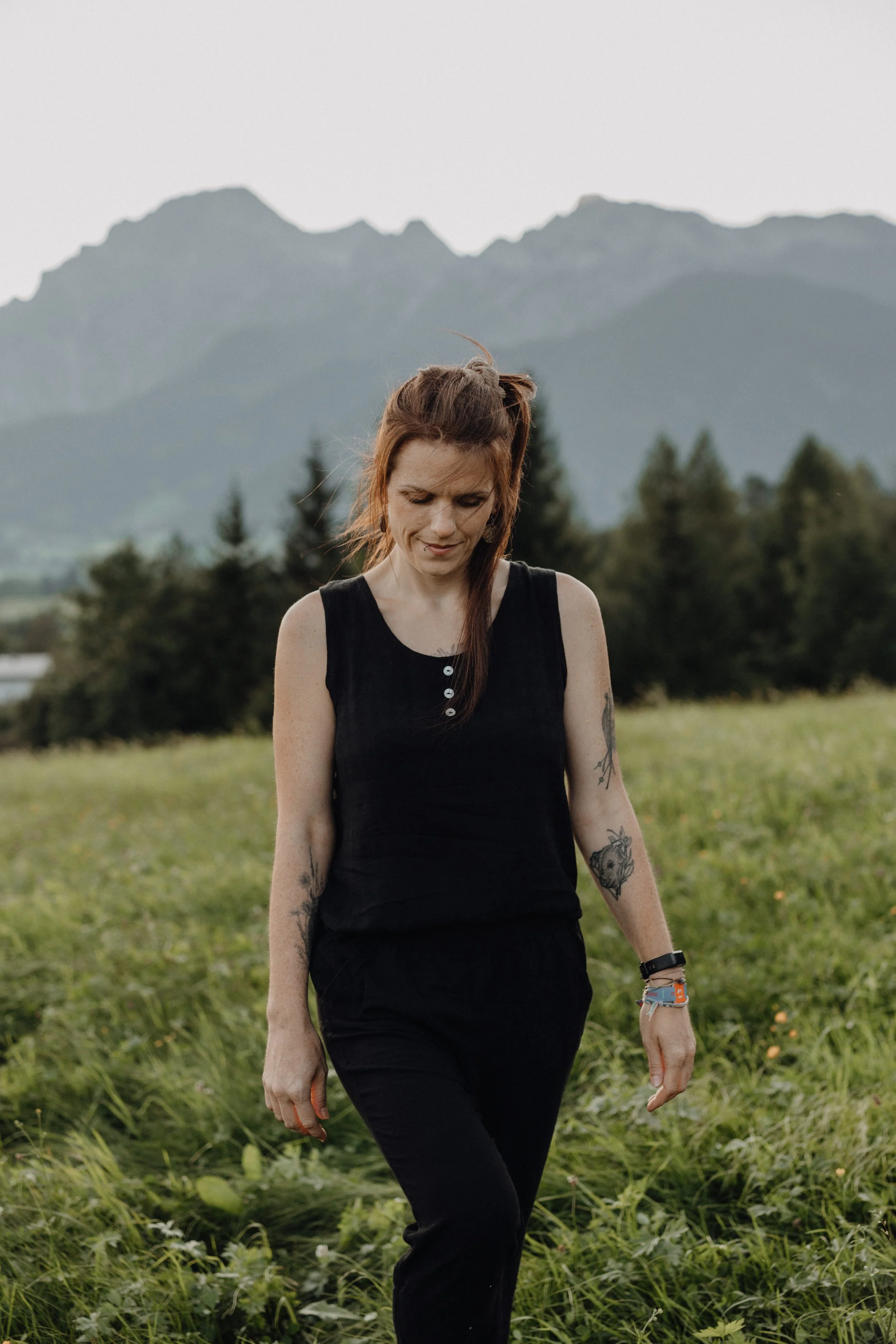 Junge Frau in schwarzen Kleidung und Tattoos, Spaziergang im Grünen mit Bergen im Hintergrund.
