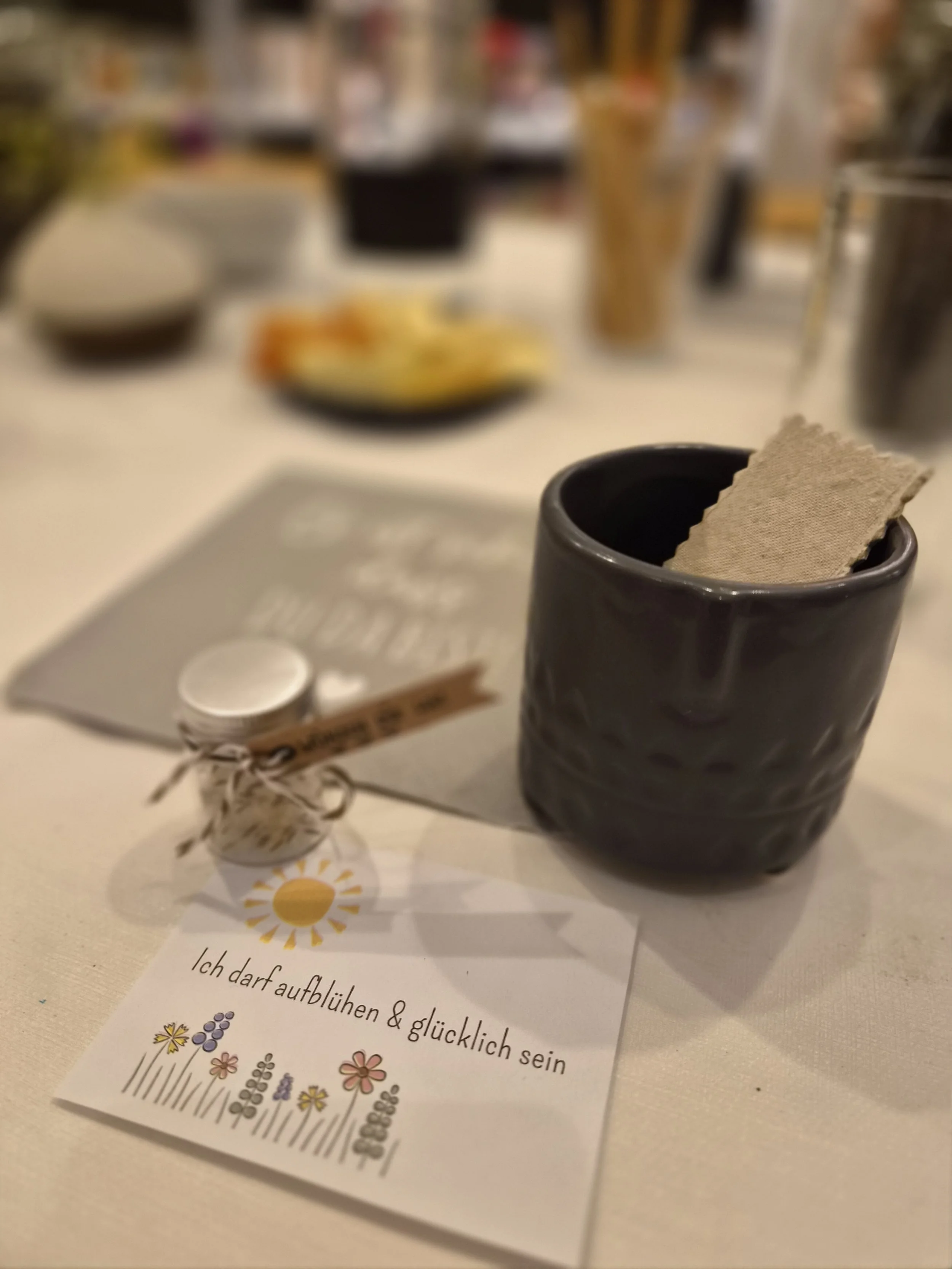 Ein schwarzer, kleiner Blumentopf steht auf einem weißen Tisch. Im Hintergrund befinden sich Blümchen-Bastelmaterialien und eine Karte mit Sonnen- und Blumenmotiven sowie dem deutschen Text "Ich darf aufblühen & glücklich sein".
