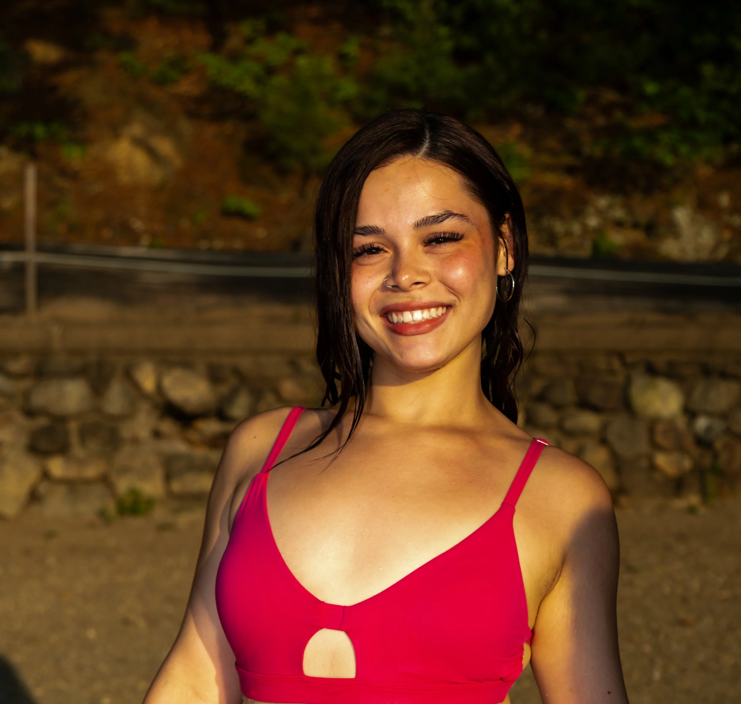 Close-up of a smiling woman with wet dark hair wearing a pink bikini top outdoors at night.