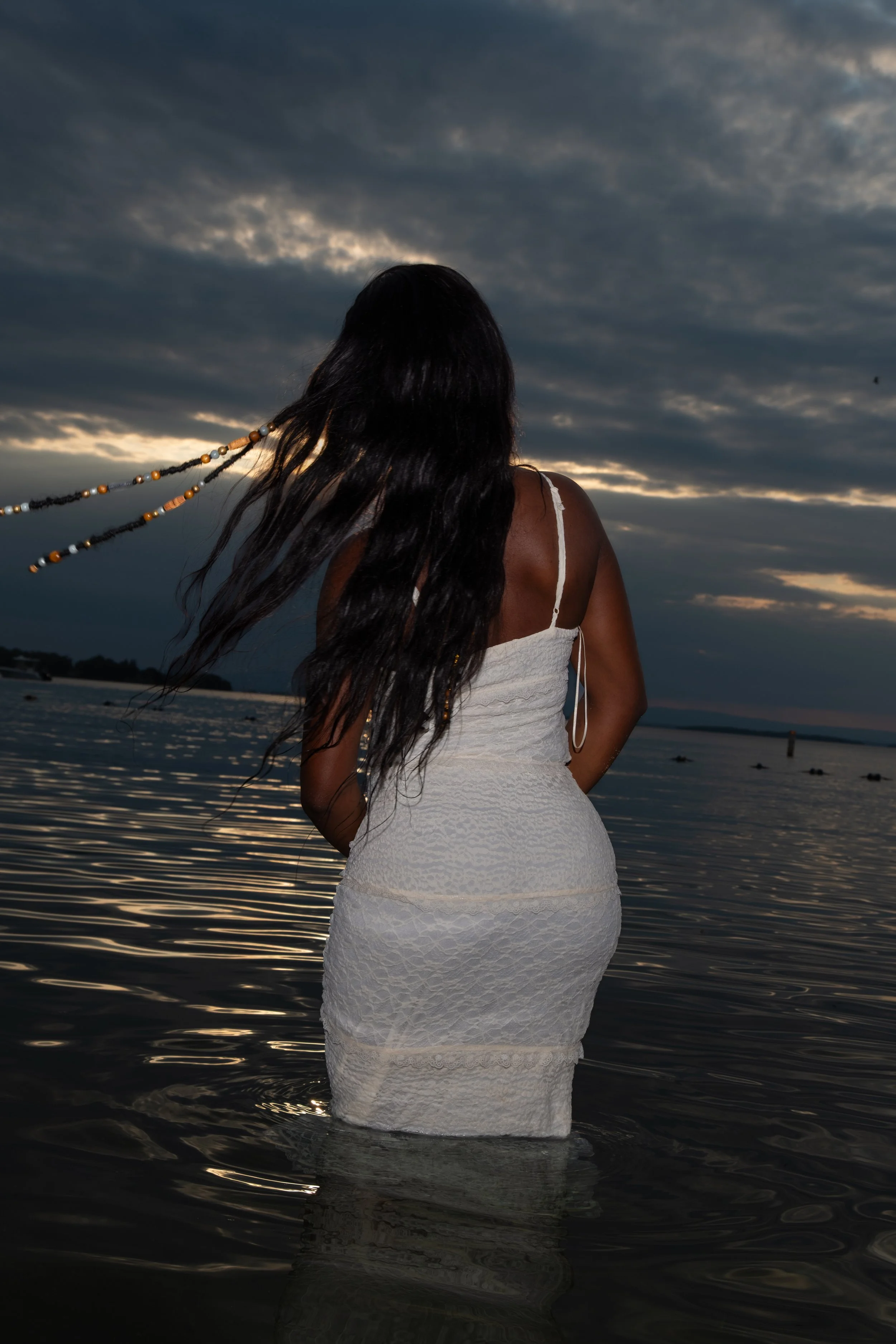 A woman in a white dress stands in the water at the beach during sunset, facing away from the camera with her long dark hair blowing in the wind.