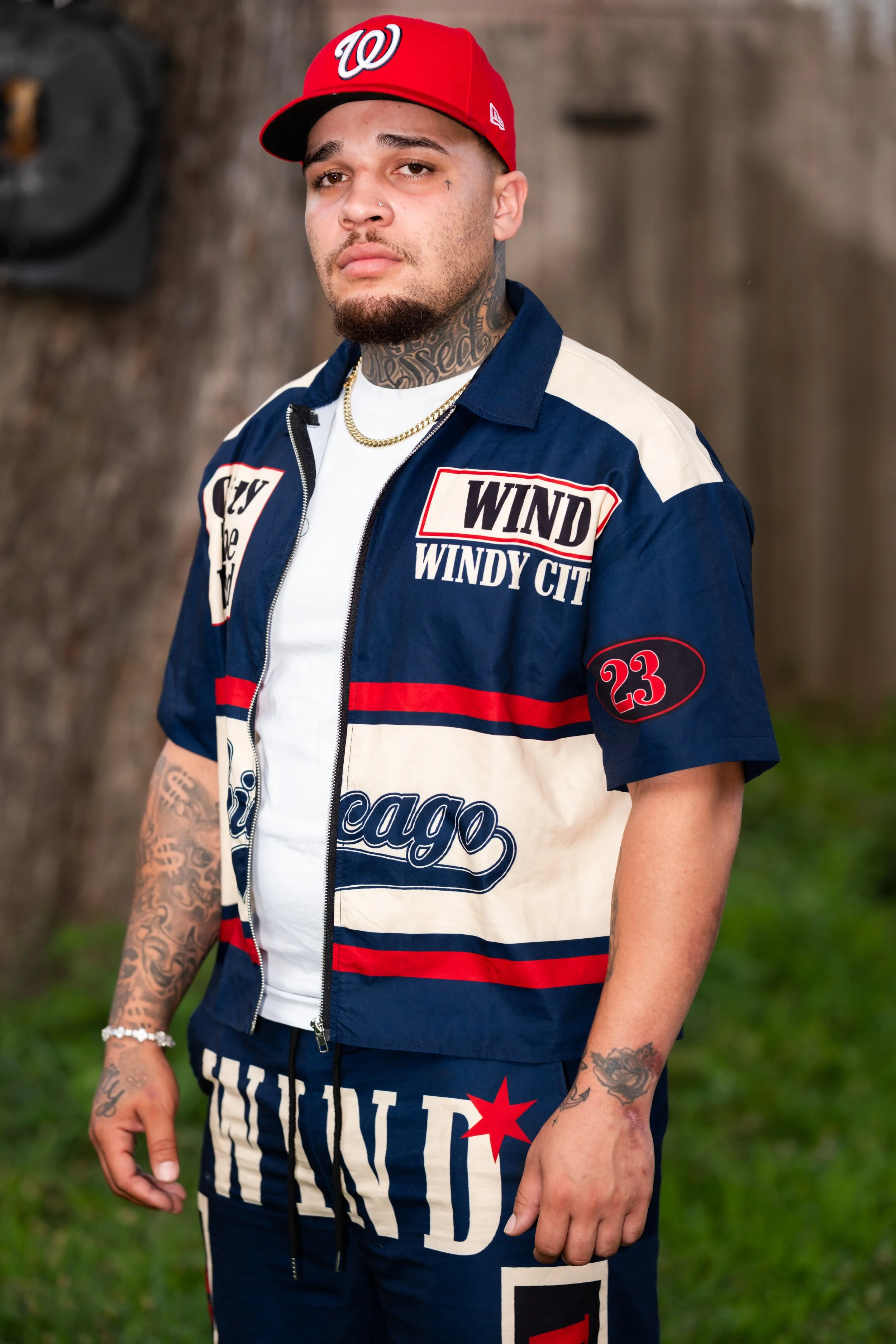 Man wearing a red Washington baseball cap, a navy and white windbreaker with patches that say "Wind" and "Windy City," and dark shorts with a Chicago logo. He has tattoos on his neck and arms, a gold chain, and a small nose piercing, standing outdoor
