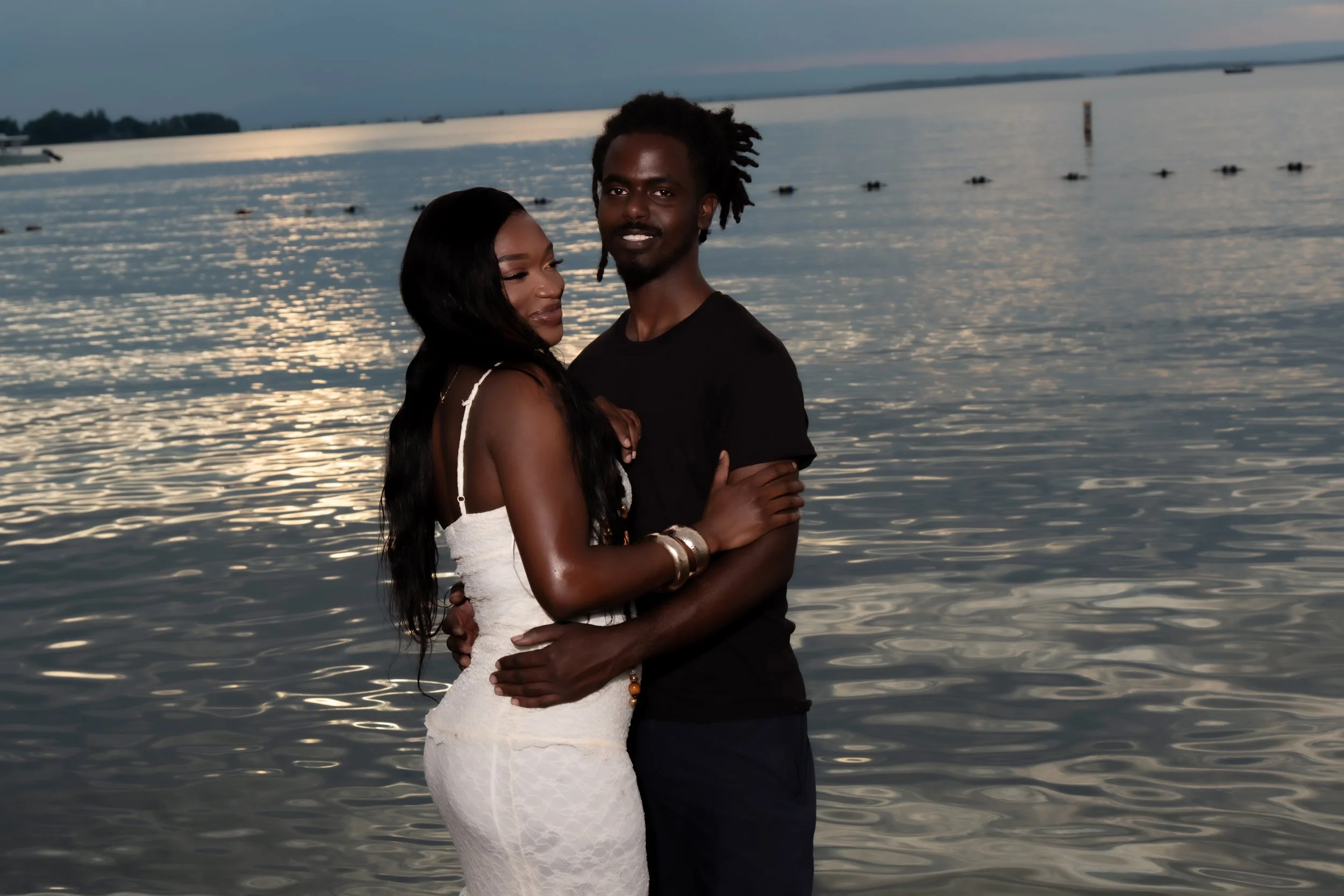 A couple standing close together by a body of water during sunset, with calm water and distant land or islands in the background. The woman is wearing a white dress and the man a black shirt, both embracing each other and smiling.