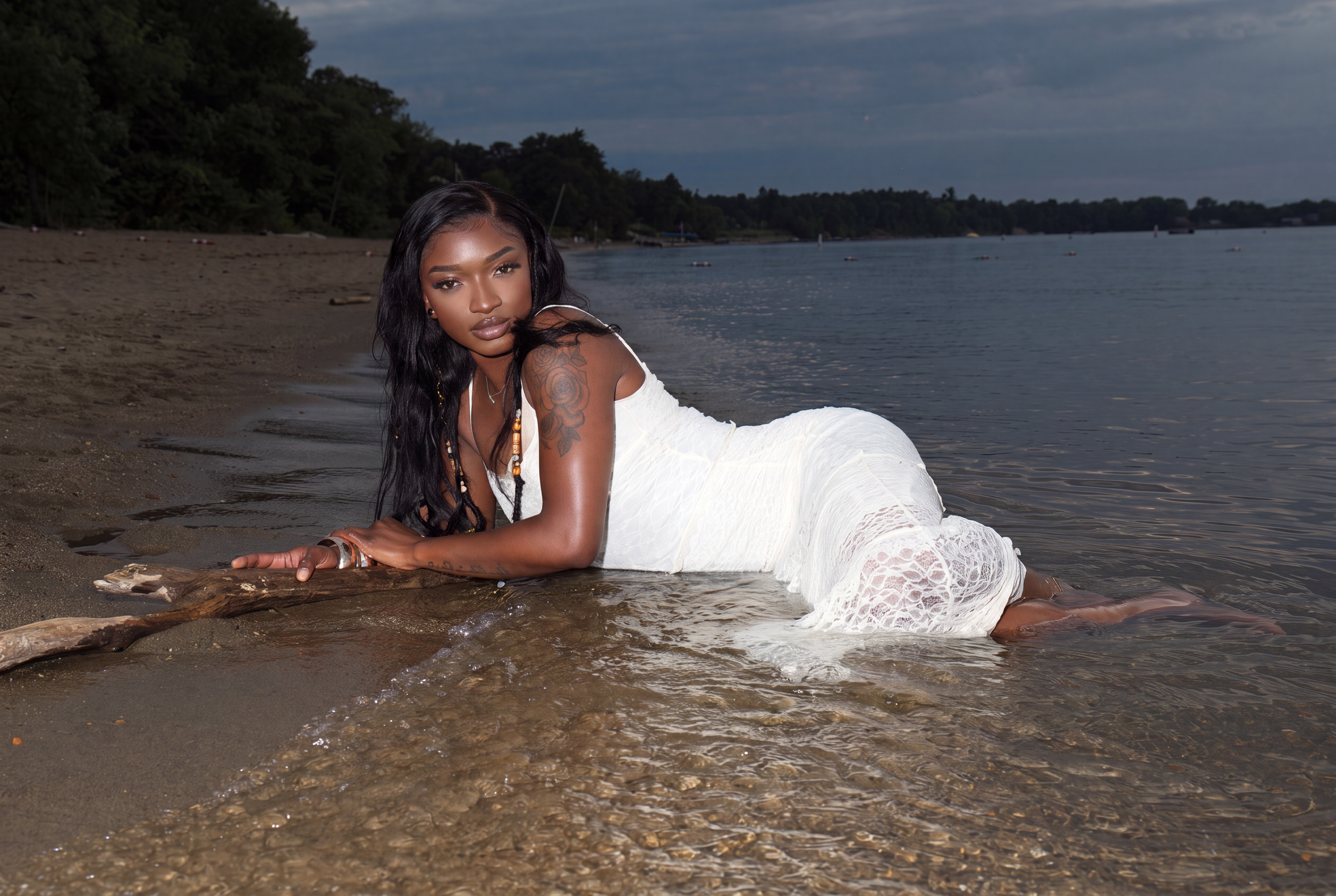 A woman with long dark hair, wearing a white lace dress and jewelry, lying on a log at a beach with water and trees in the background.