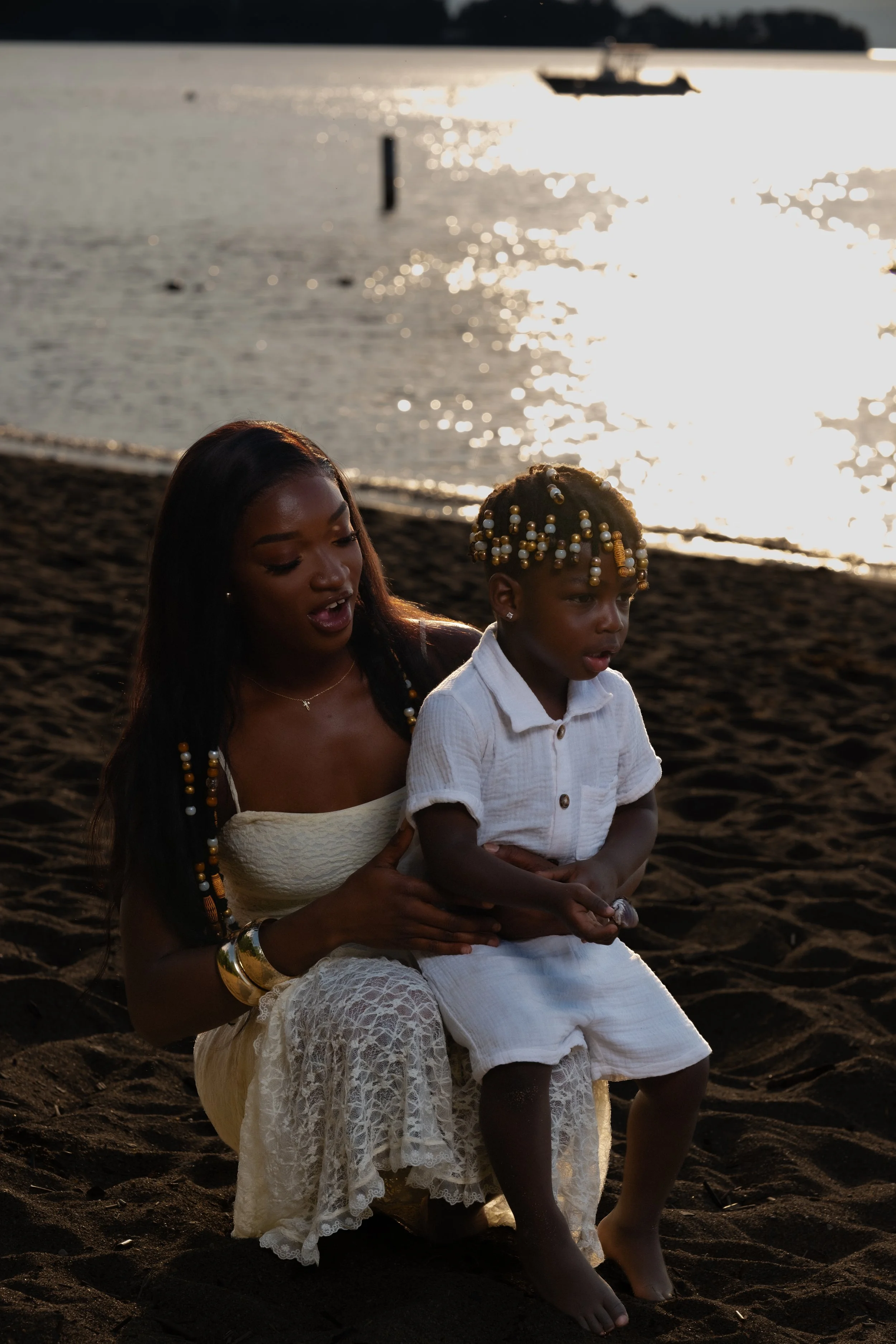 A woman and a young girl sitting on the sandy beach at sunset, with a boat and water in the background.