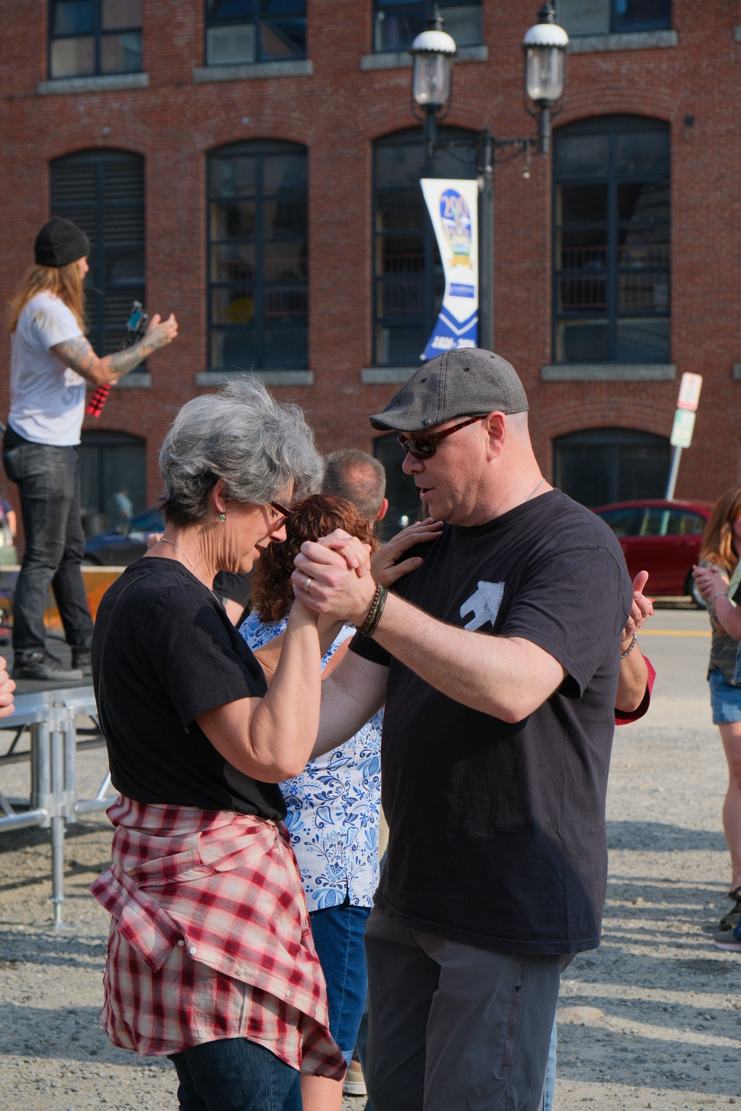An elderly woman with gray hair, glasses, wearing a black shirt, and a red plaid shirt tied around her waist, dancing with a middle-aged man with a gray hat, sunglasses, and a black T-shirt during an outdoor event. Other people and a red brick buildi
