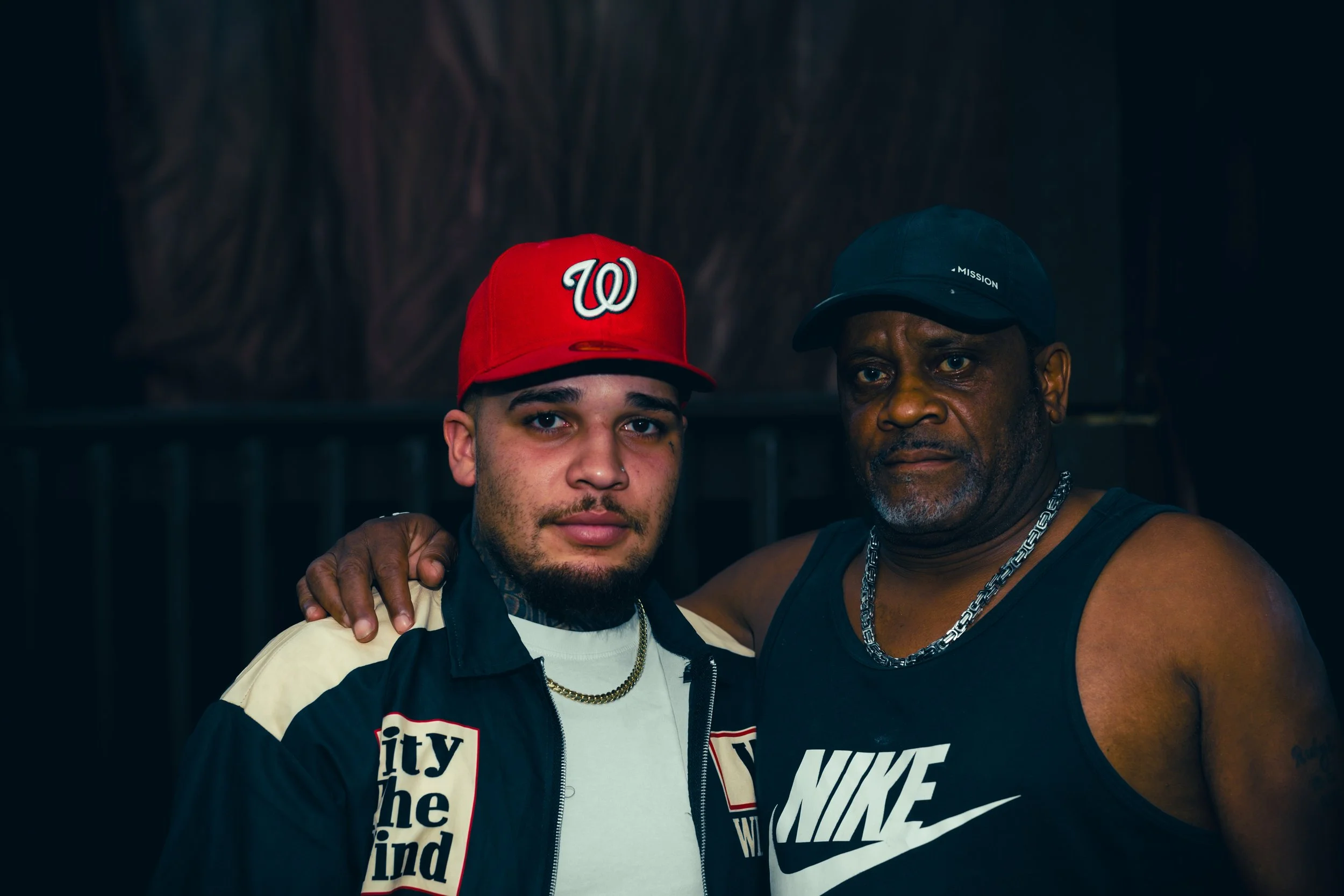 Two men posing together; one wearing a red baseball cap with a Washington Wizards logo, a cream and black jacket, and a chain necklace; the other wearing a black cap with 'MISSION' written on it, a black tank top, and a chain necklace. They are smili